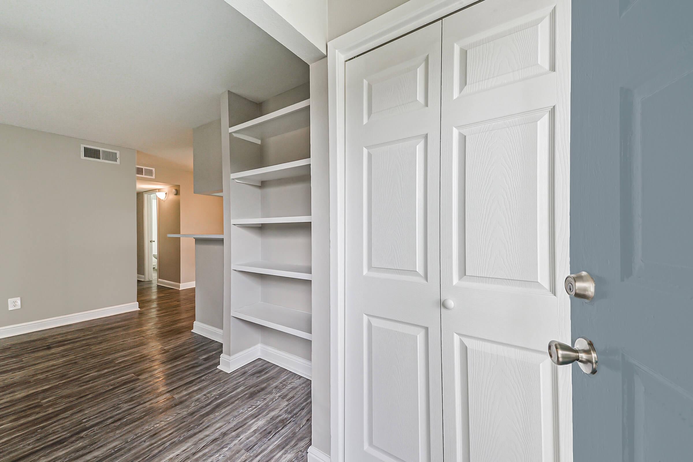 A hallway leading into a room with light-colored walls and hardwood flooring. On the left, there is a white double-door closet, and to the right, open shelving. The background shows another room, illuminated by natural light, suggesting a spacious layout.