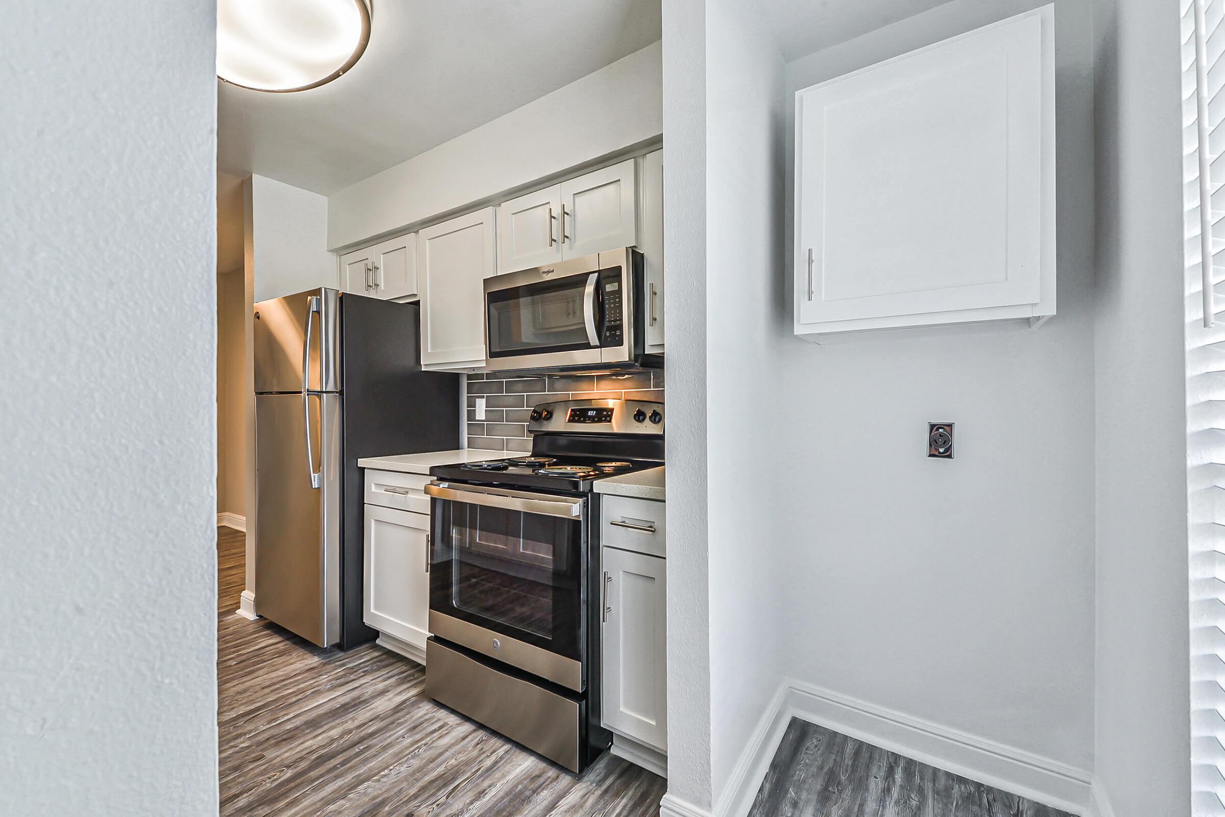 A modern kitchen featuring white cabinetry, stainless steel appliances including a refrigerator and gas stove, and a tile backsplash. The space has a neutral color scheme with wood-like flooring and a small pantry or storage cupboard. Natural light streams in from a nearby window.