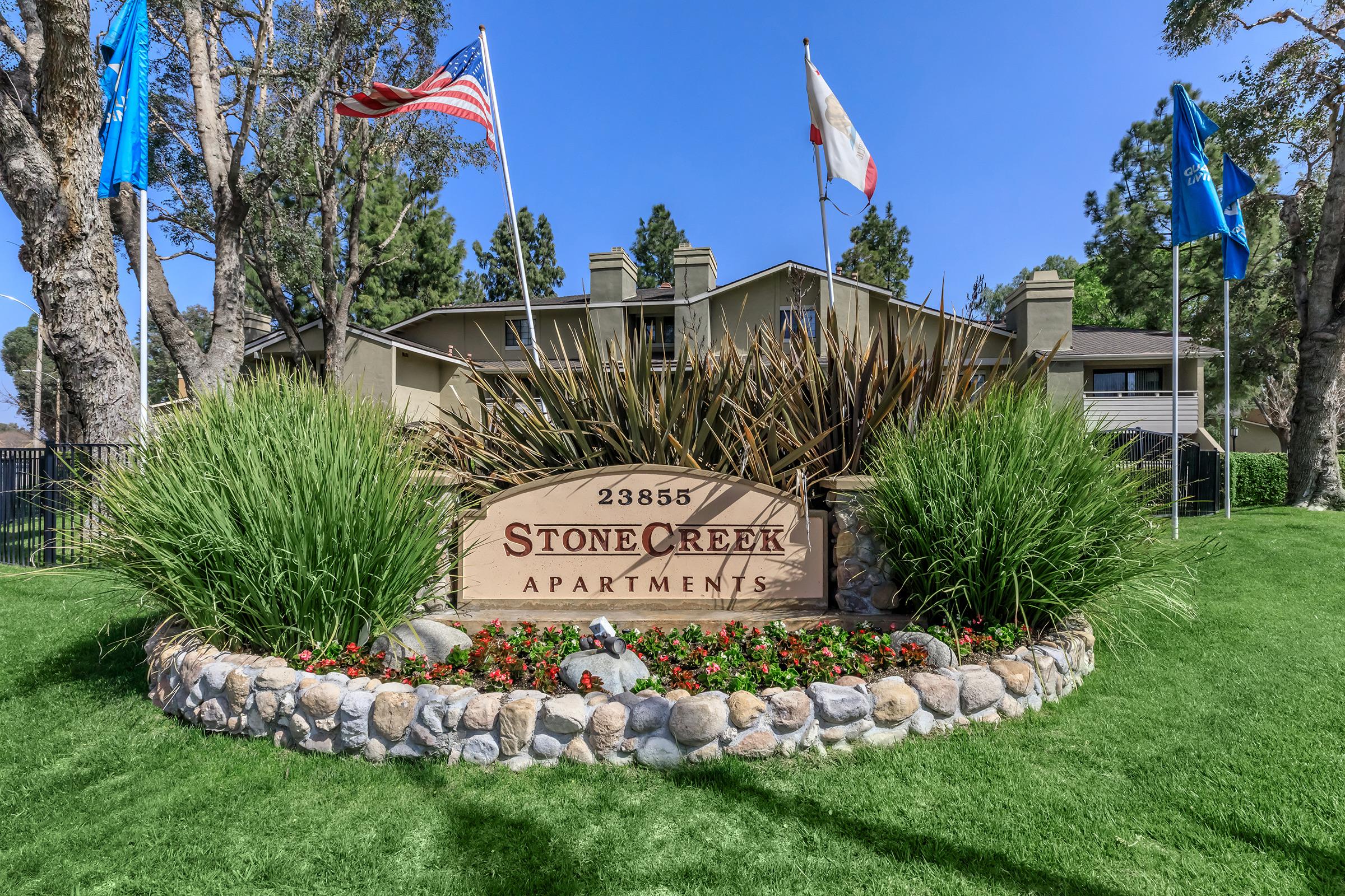 A sign for Stone Creek Apartments, featuring the address 23855, surrounded by decorative plants and flowers. The sign is flanked by multiple flags and is set against a clear blue sky and green grass. The apartment complex is visible in the background.