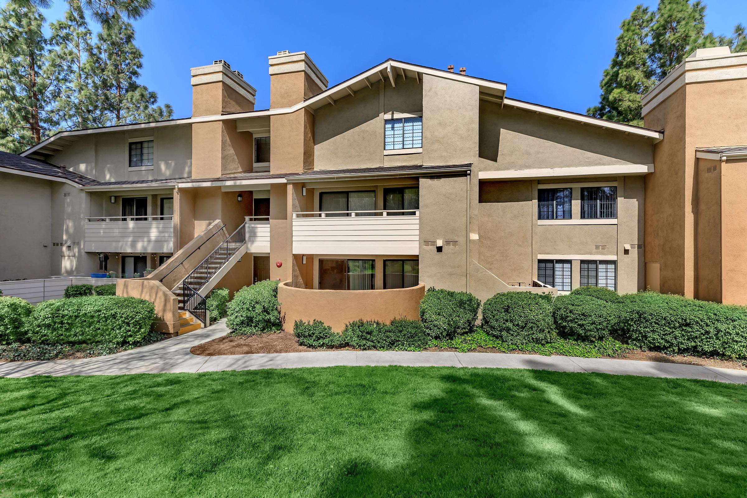 Two-story apartment building with a stucco exterior in light brown and cream tones. The building features balconies, large windows, and a well-maintained lawn with shrubs in the foreground. Stairs lead to the second level on one side, and the environment appears sunny and inviting, surrounded by trees.