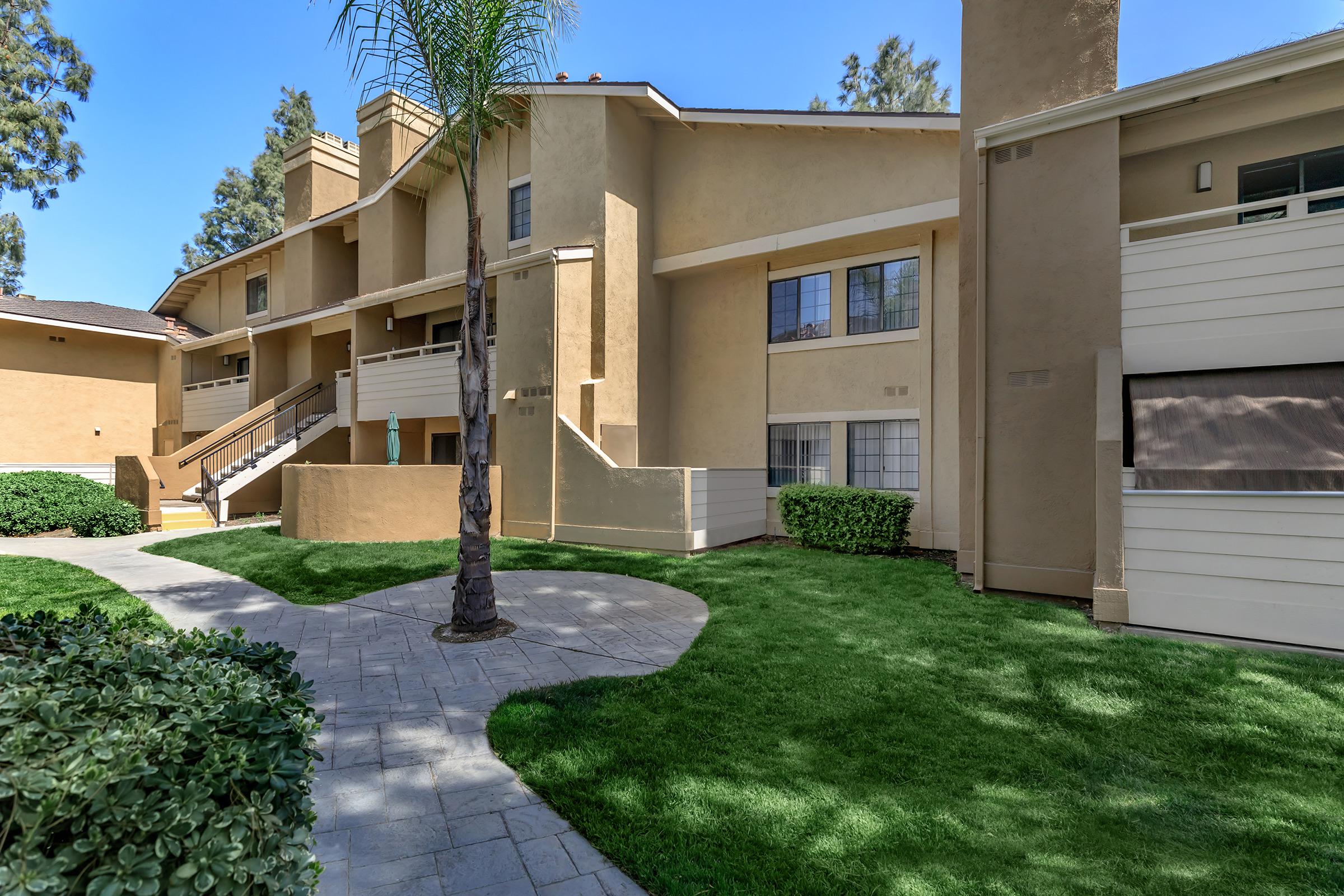 A well-maintained apartment complex featuring two-story buildings with beige exteriors, surrounded by lush green lawns and shrubs. A paved pathway winds through the landscaping, and a palm tree adds a tropical touch to the scene. Bright blue sky is visible overhead, indicating clear weather.
