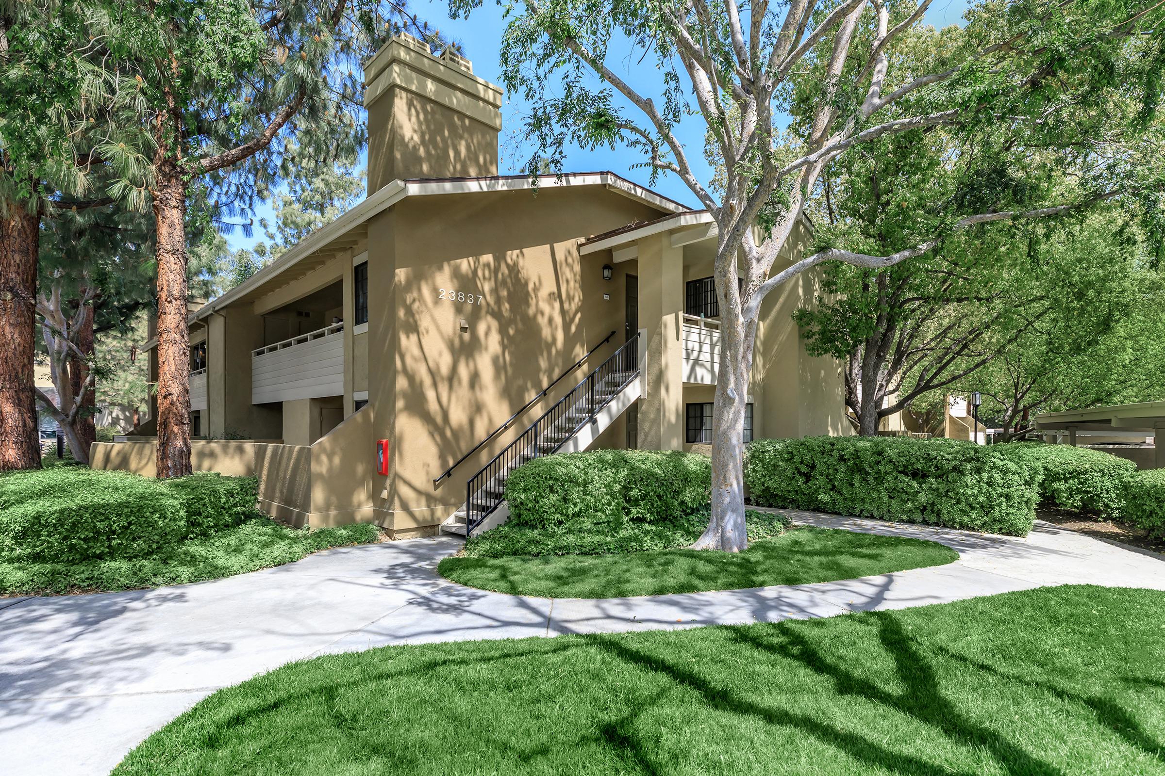 A multi-story apartment building surrounded by lush greenery and well-maintained landscaping. The structure features a light beige exterior with balconies, and a winding pathway leading through manicured lawns and trees. Sunlight filters through the leaves, creating a pleasant and inviting atmosphere.