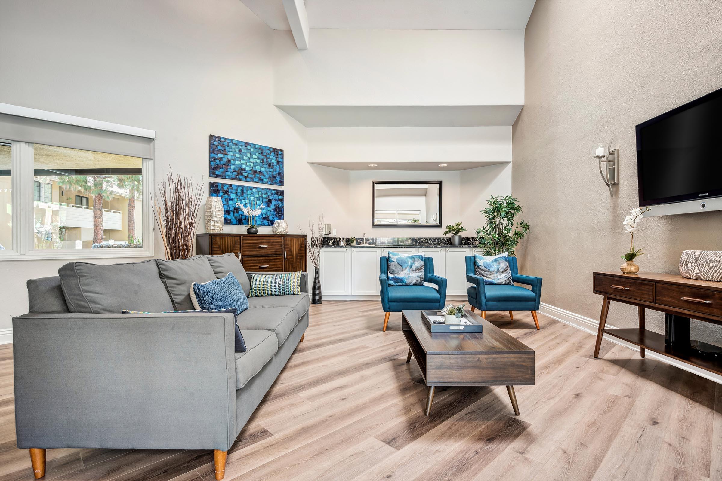 A modern living room featuring a gray sectional sofa with decorative pillows, two blue accent chairs, a wooden coffee table, and a wall-mounted TV. The space is adorned with abstract wall art, indoor plants, and natural light from large windows, creating a warm and inviting atmosphere.