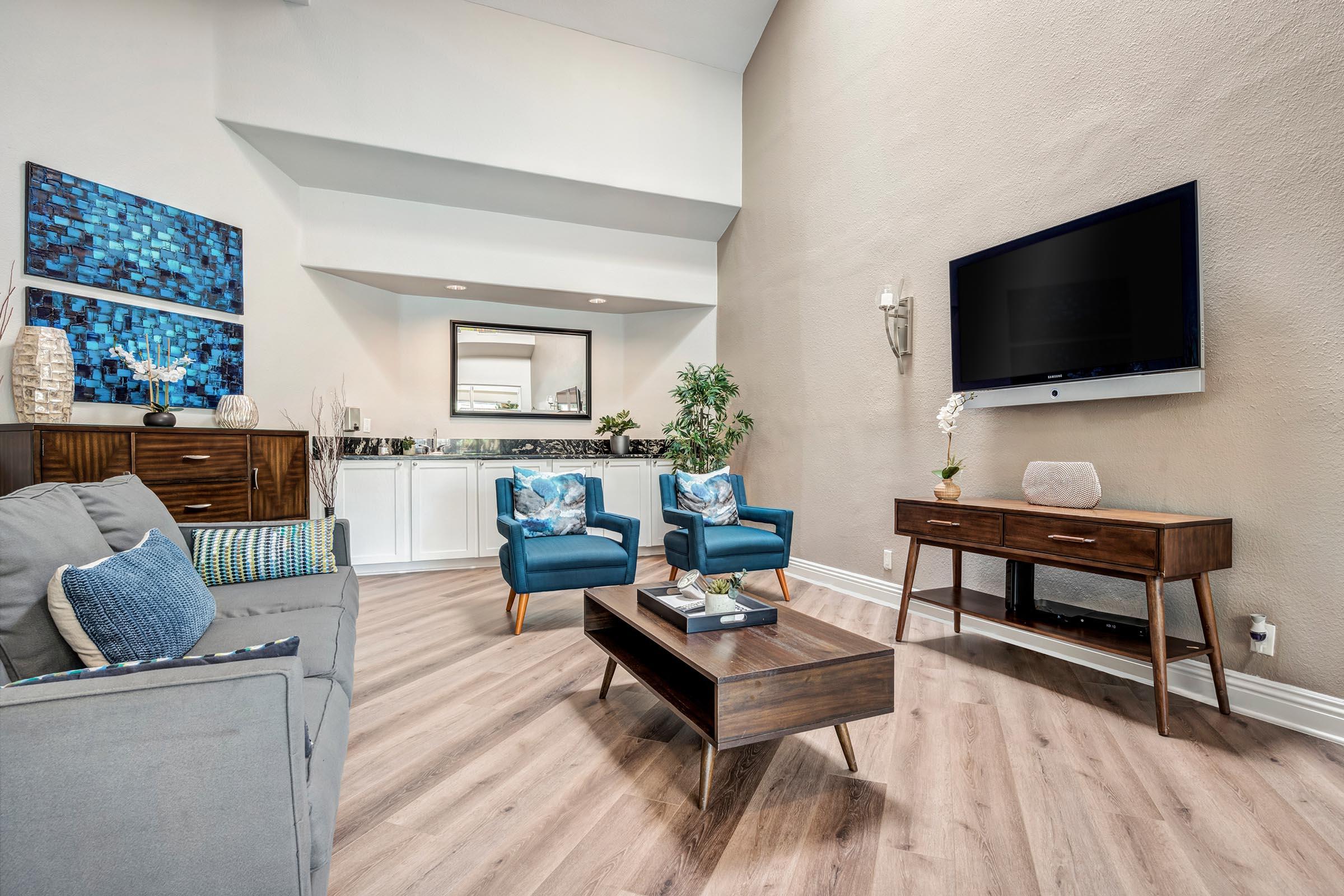 A modern living room featuring a gray sofa, two blue armchairs, and a wooden coffee table. A large flat-screen TV is mounted on the wall, and there are decorative plants and artwork in the background. The room has light-colored walls and wooden flooring, creating a bright and inviting atmosphere.
