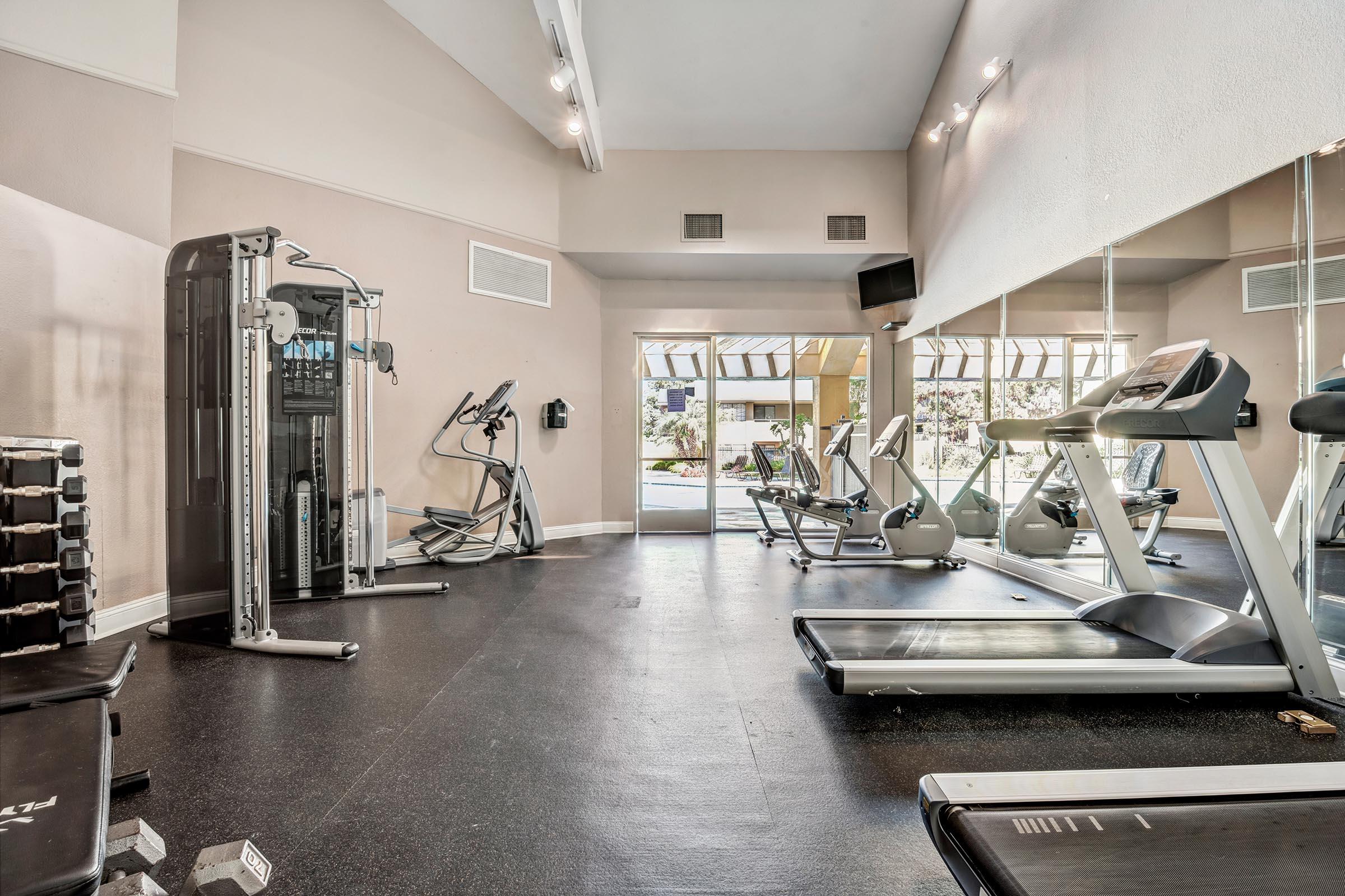 A well-lit gym featuring various exercise equipment, including treadmills, an elliptical machine, a weight machine, and free weights. Large mirrors line one wall, and large windows allow natural light to fill the space, creating an inviting atmosphere for workouts.