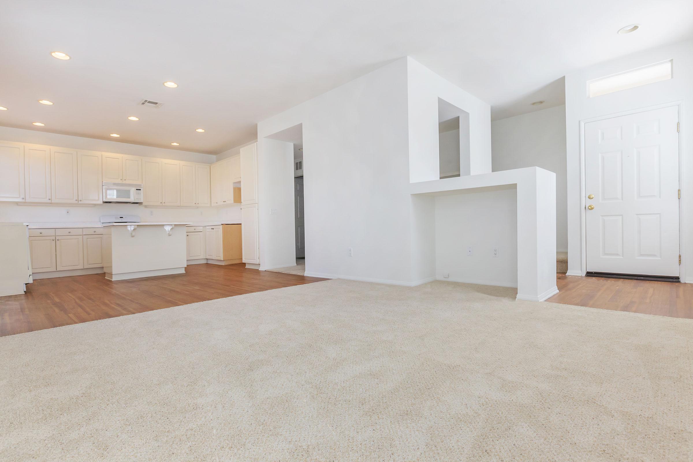 A spacious, empty living area featuring light-colored walls and beige carpet. The room has a doorway leading outside, alongside an open kitchen with light cabinetry and modern appliances. Bright overhead lights illuminate the space, creating a welcoming atmosphere.