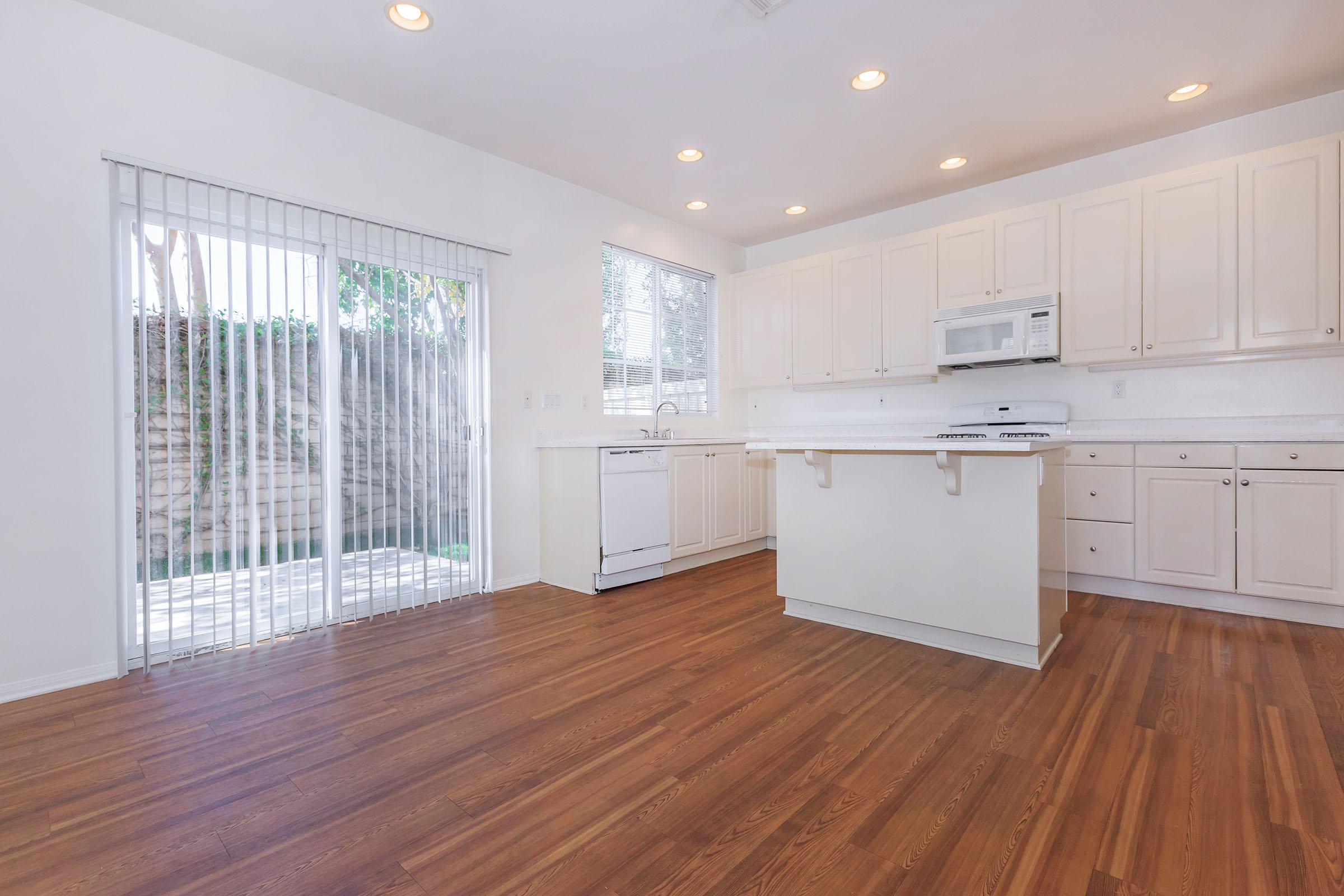 A bright, modern kitchen featuring white cabinetry, a microwave, and a dishwasher. The space has wooden flooring and large windows with blinds that let in natural light. A sliding glass door provides access to an outdoor area, enhancing the open, airy feel of the room.