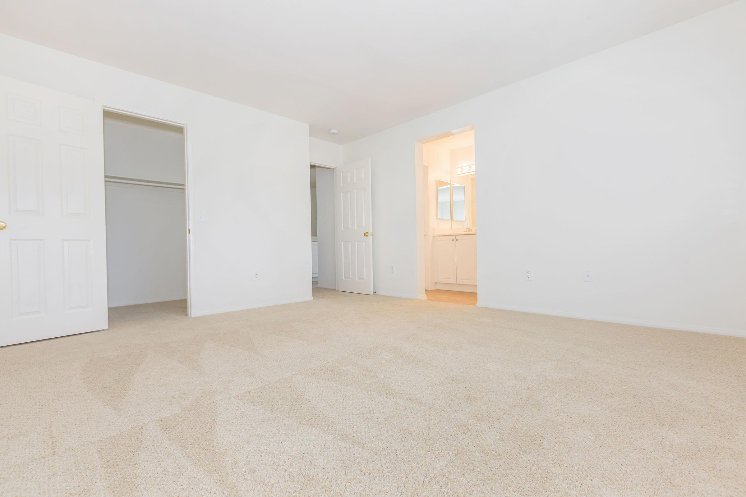 An empty room with light beige carpet, white walls, and two doors leading to adjacent spaces. One door opens to a closet, and another leads to a well-lit bathroom or hallway area. The space is bright and uncluttered, providing a blank canvas for decoration.