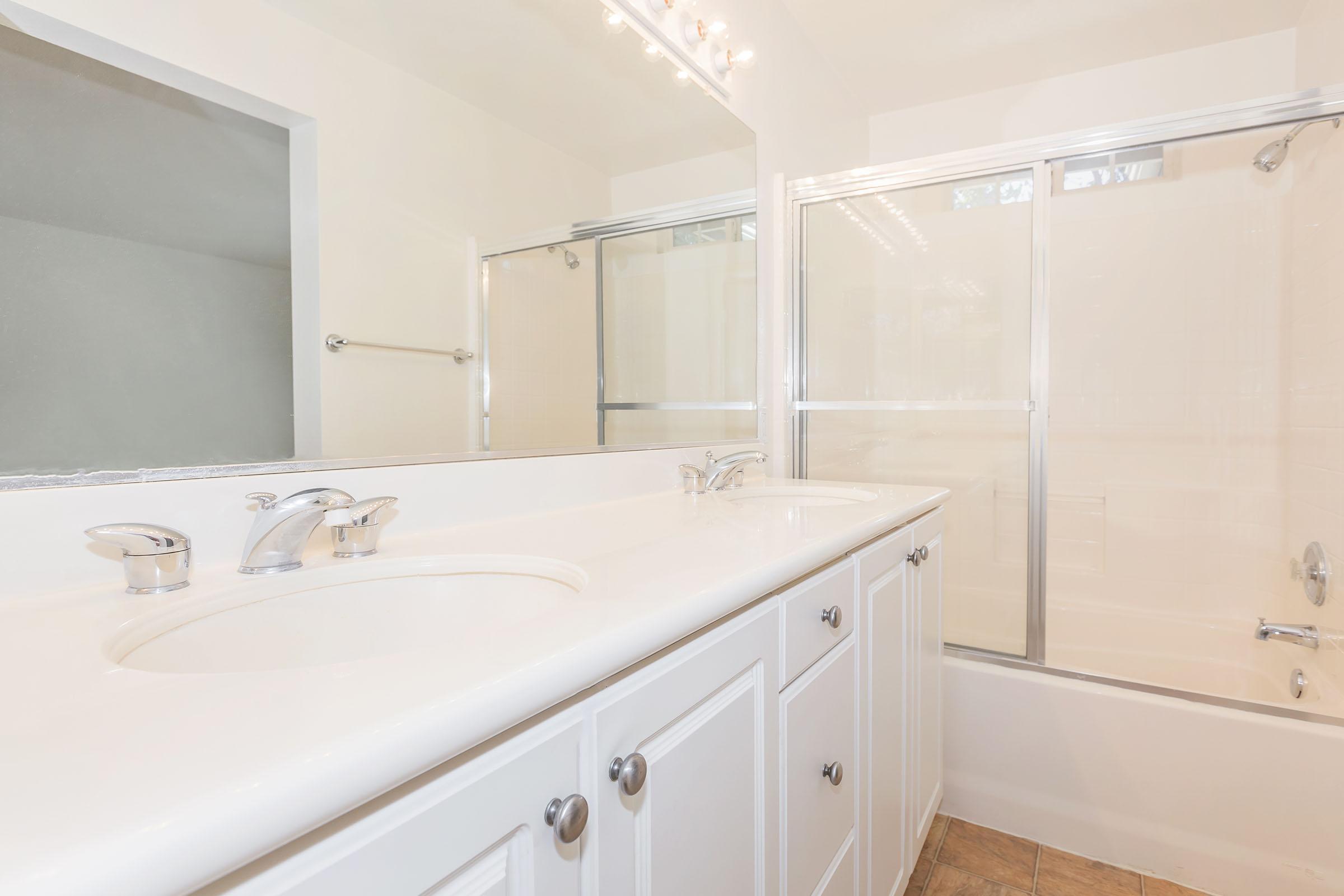 A bright and clean bathroom featuring a double sink vanity with silver faucets, a large mirror above the sink, and a glass shower enclosure. The walls are light-colored, and there is a bathtub with a showerhead. Neutral-toned flooring completes the spacious, modern look of the bathroom.
