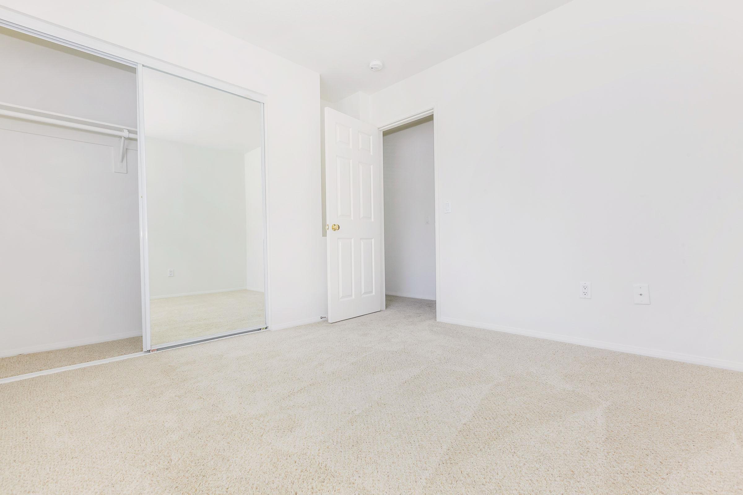 Empty room with beige carpet, white walls, and a large mirror closet on the left. A closed door leads to another room, positioned slightly to the right of the image. The space is bright and uncluttered, creating a minimalist atmosphere.
