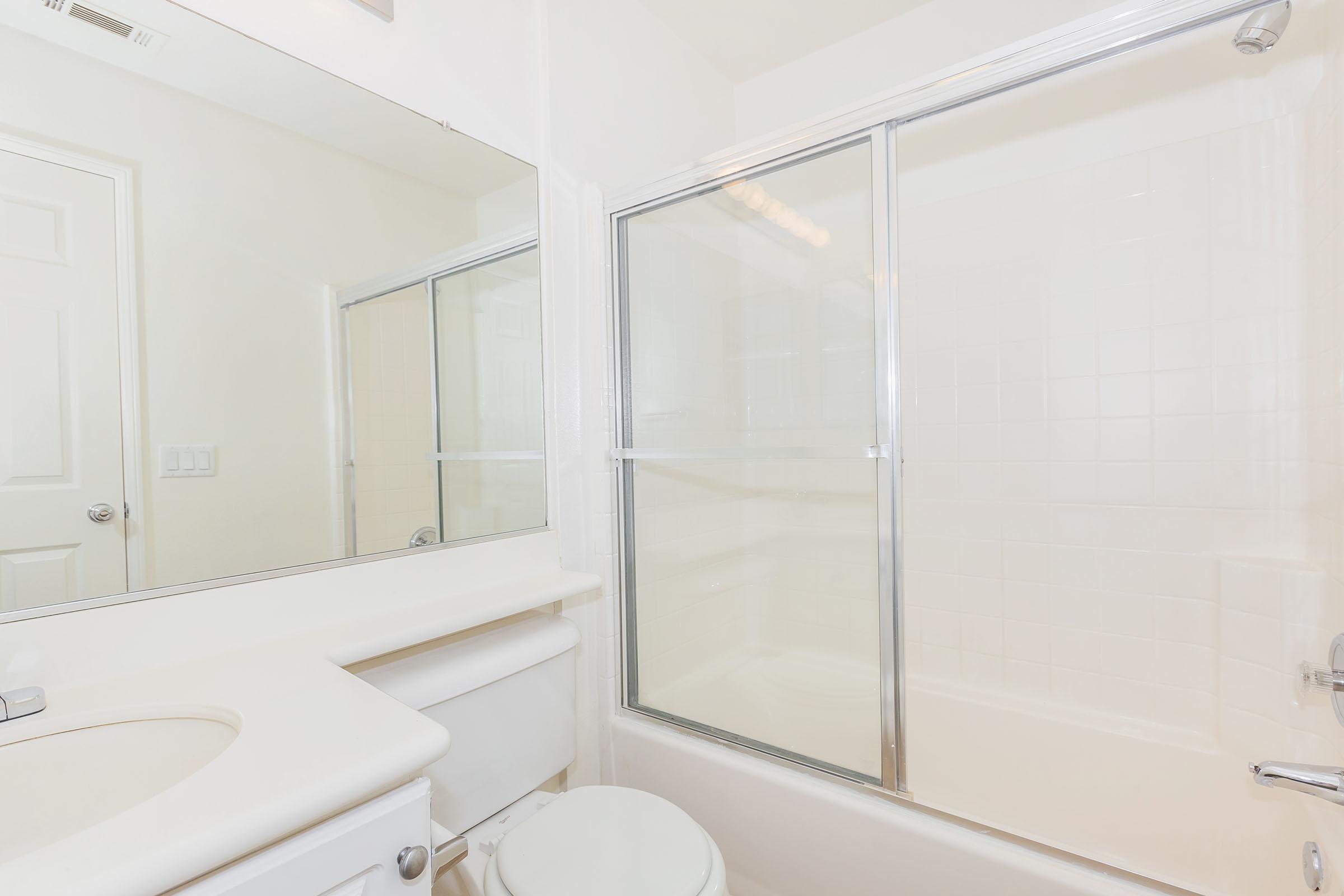 A clean, well-lit bathroom featuring a white bathtub with a clear glass door, a rectangular mirror above the sink, and white walls. The sink is positioned next to the bathtub, and there is a door leading to another area of the space. The overall aesthetic is modern and minimalist.