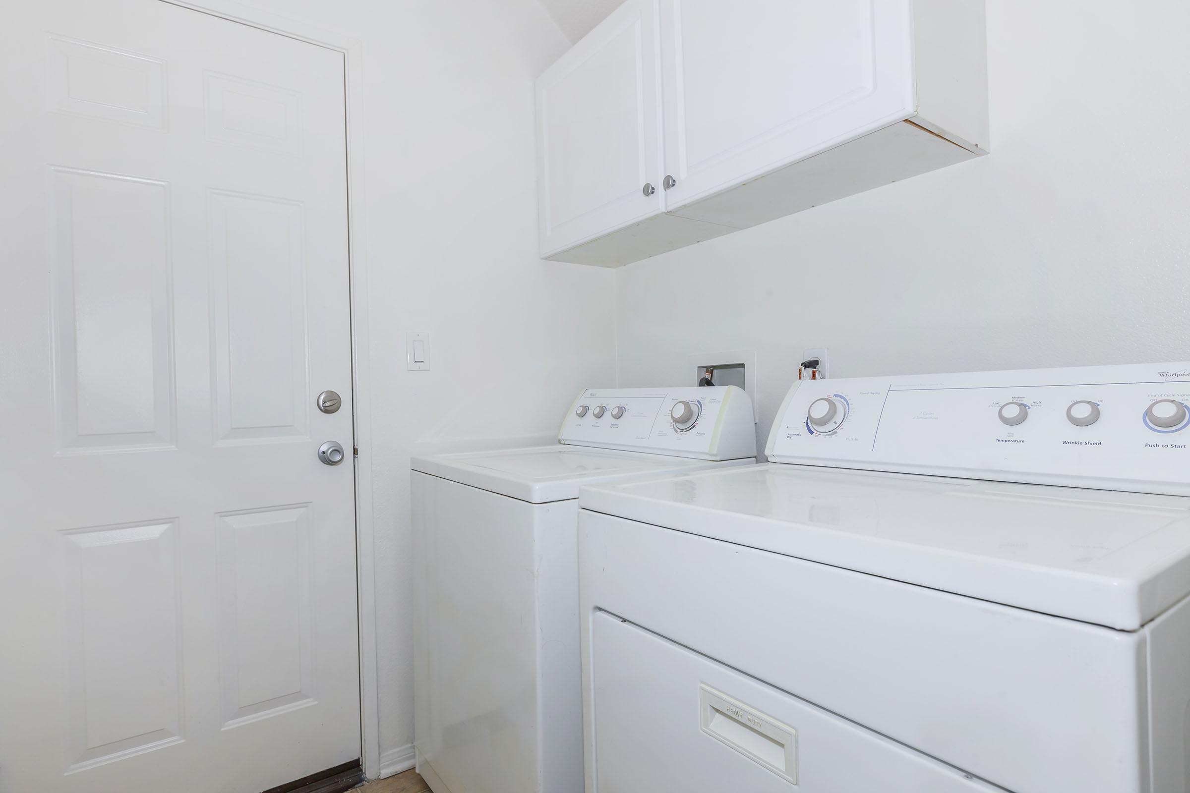 A clean laundry room featuring a white washing machine and dryer side by side. The walls are painted white, and there is a simple white door visible. The floor has light-colored wooden panels, creating a bright and airy atmosphere in the space.