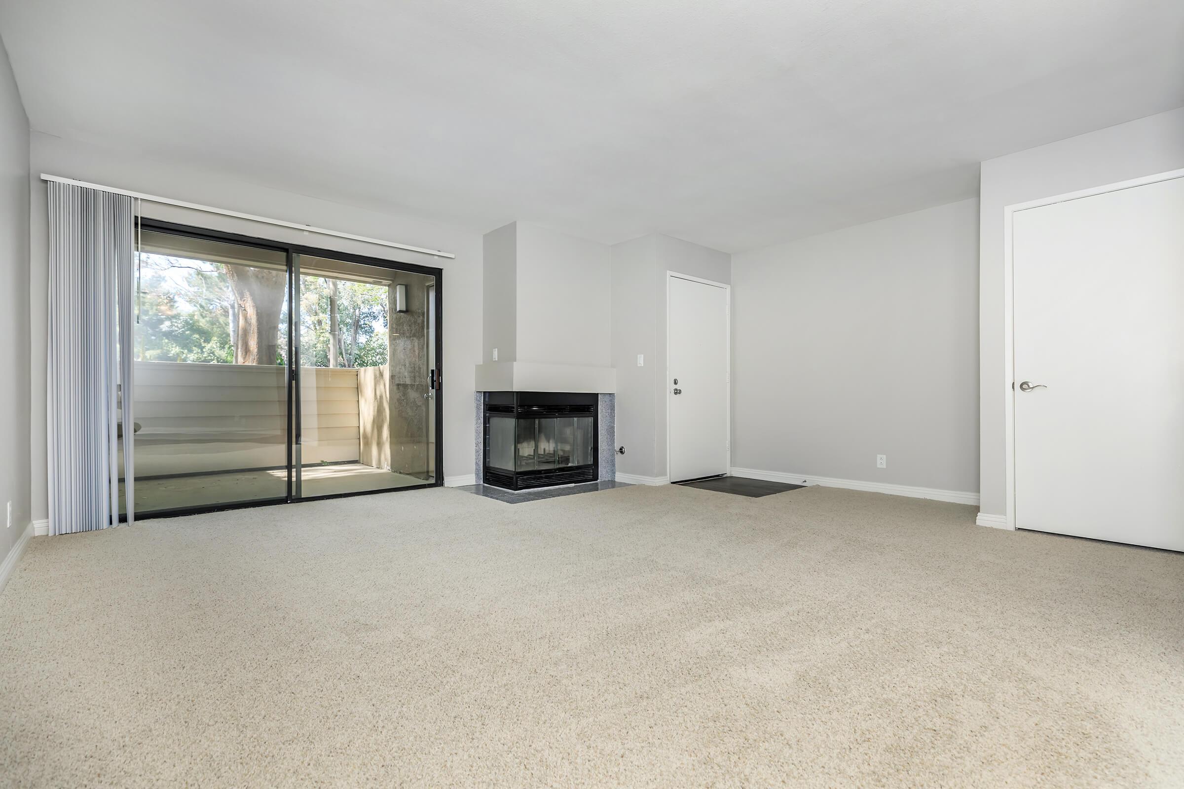 A spacious, empty living room with light beige carpet, a fireplace with a dark stone surround, and large sliding glass doors that lead to a balcony. The walls are painted a light gray, and there are two doors visible: one to the entrance and another leading to an adjacent room. Natural light fills the space.