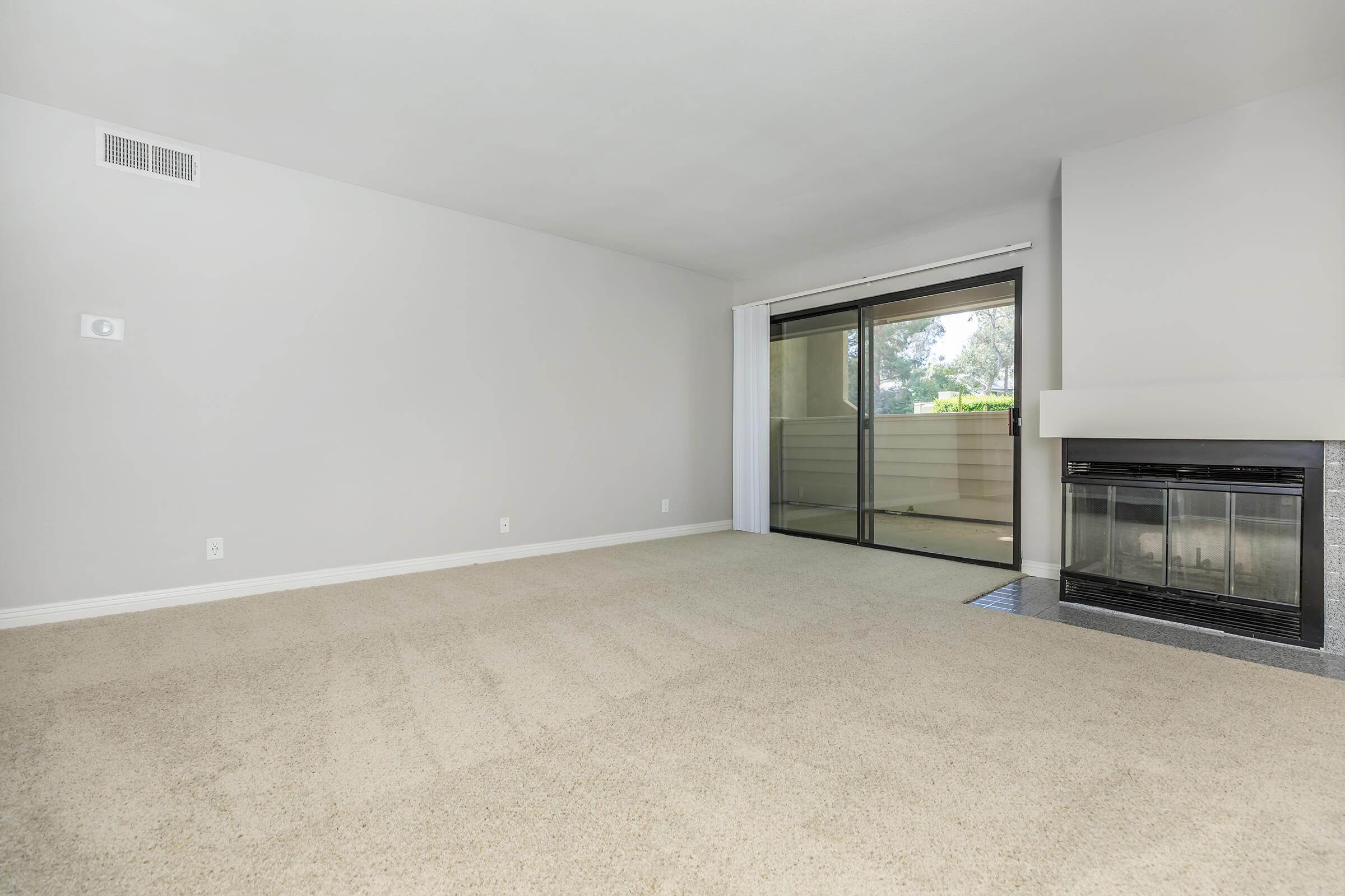 A spacious, empty living room with light gray walls and beige carpet. It features a large window with sliding glass doors leading to a balcony, and a modern fireplace on the right side of the room. The overall ambiance is bright and inviting, with ample natural light streaming in.