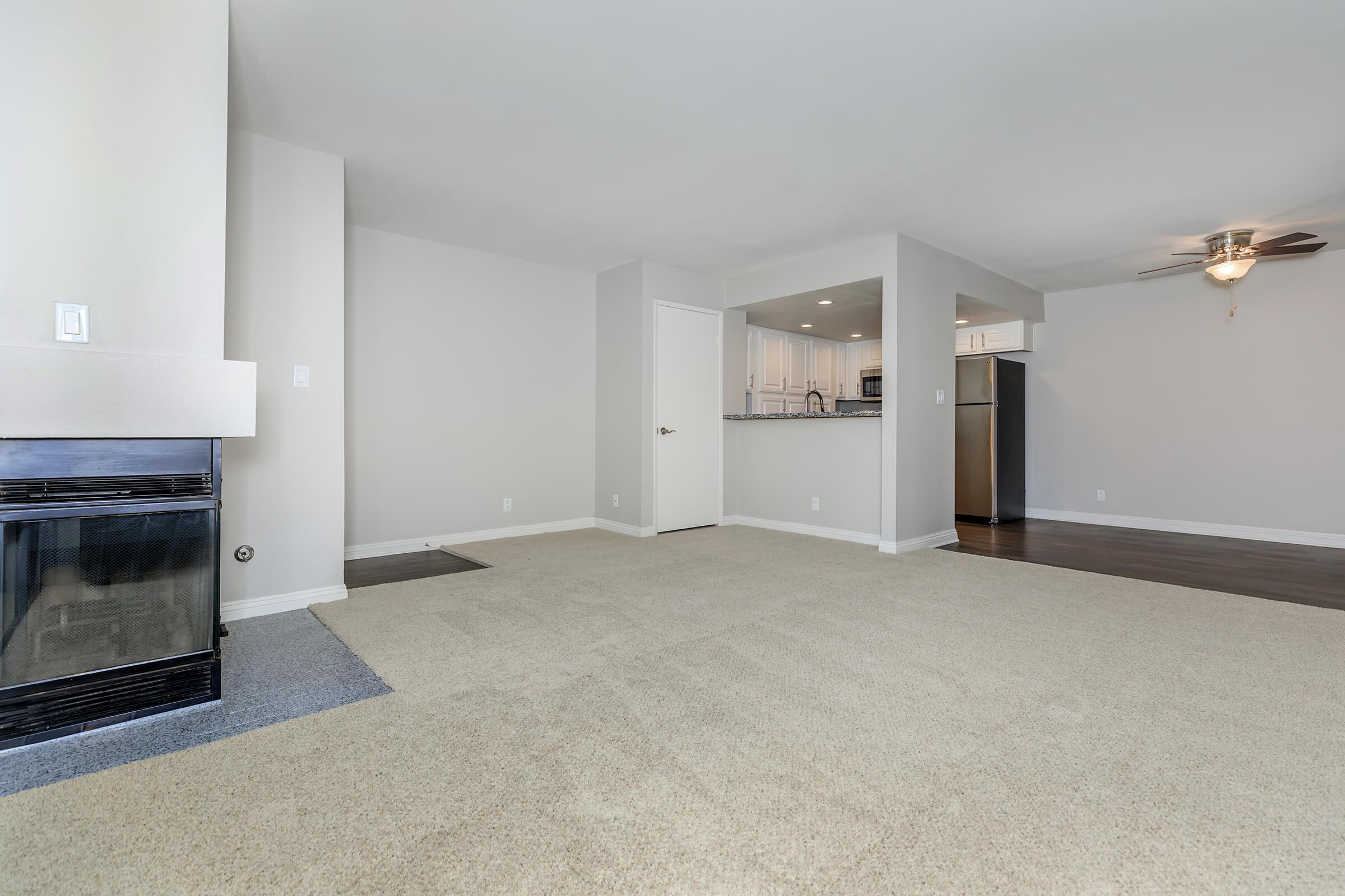 A spacious, modern living area with beige carpet and light gray walls. To the left, there's a fireplace with a stylish mantel. An opening leads to a kitchen with white cabinetry and stainless steel appliances visible in the background. A ceiling fan is mounted above, adding to the room's inviting atmosphere.