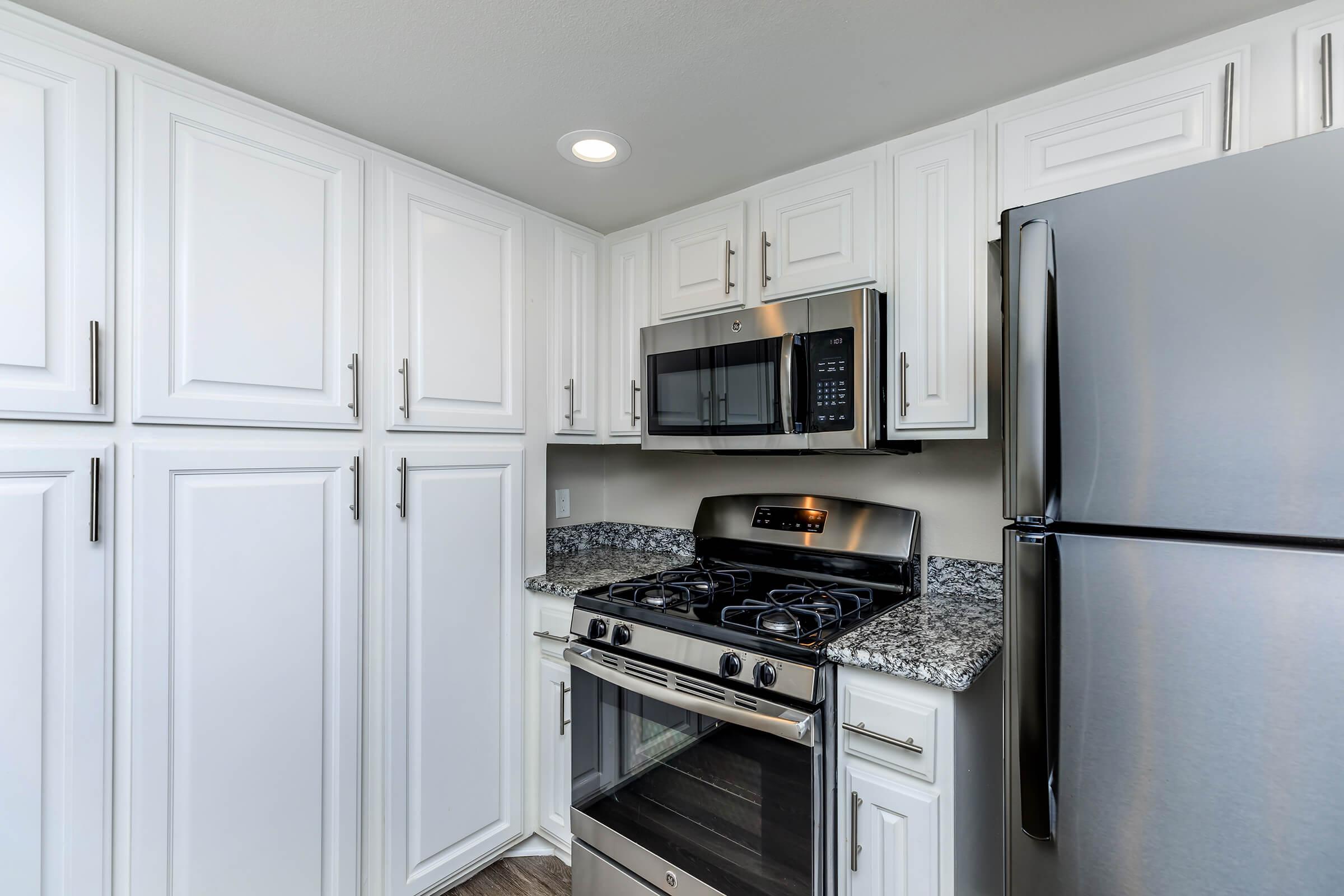 Modern kitchen featuring white cabinets, a stainless steel microwave and refrigerator, and a gas stove with a black granite countertop. The space is well-lit with overhead lighting, providing a clean and contemporary look.