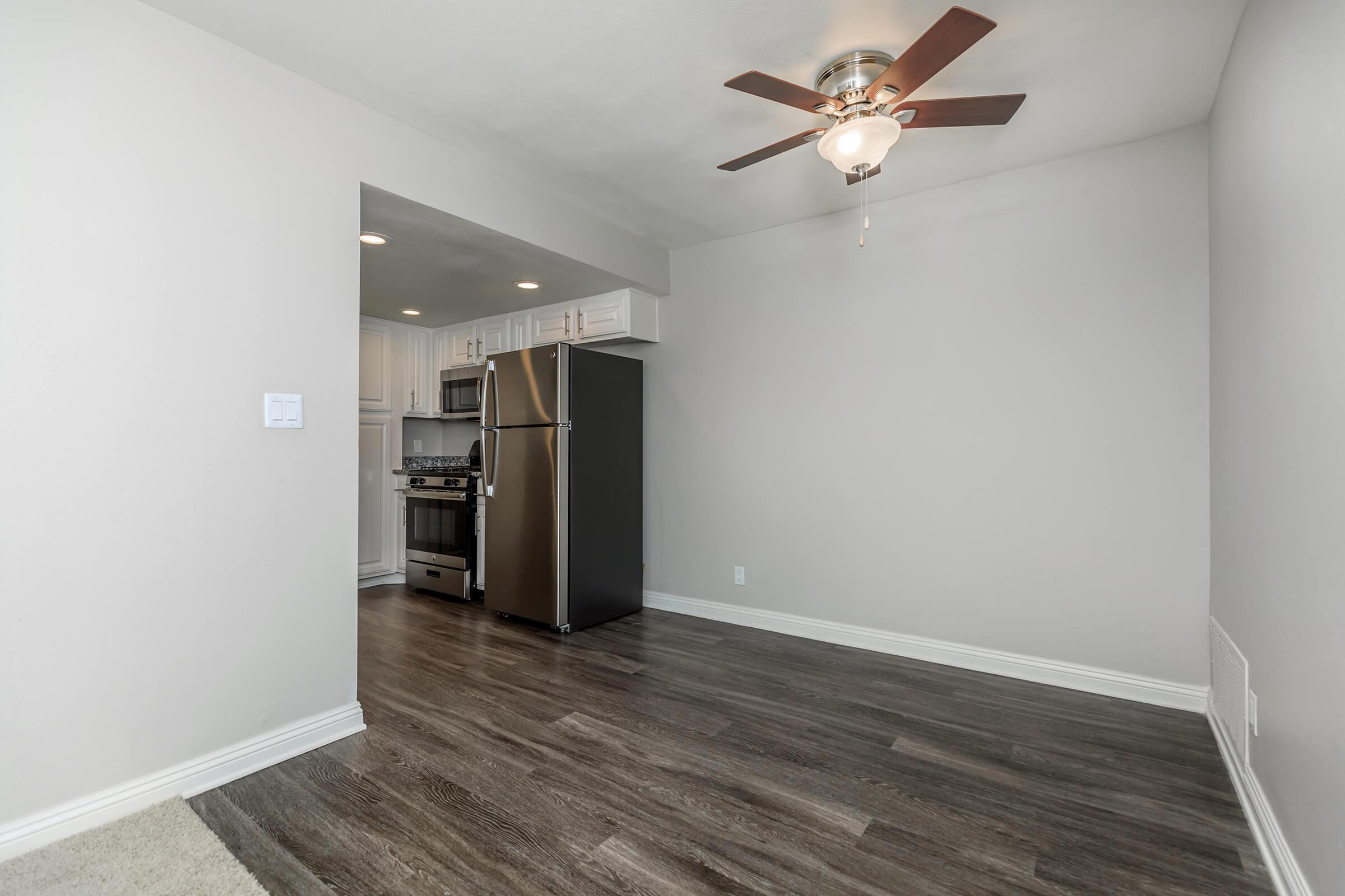 Interior of a small, modern kitchen and living space. The kitchen features stainless steel appliances, including a refrigerator and gas stove, with white cabinets above. The living area has laminate flooring and a ceiling fan. Wall is painted light gray, creating a bright and open atmosphere.
