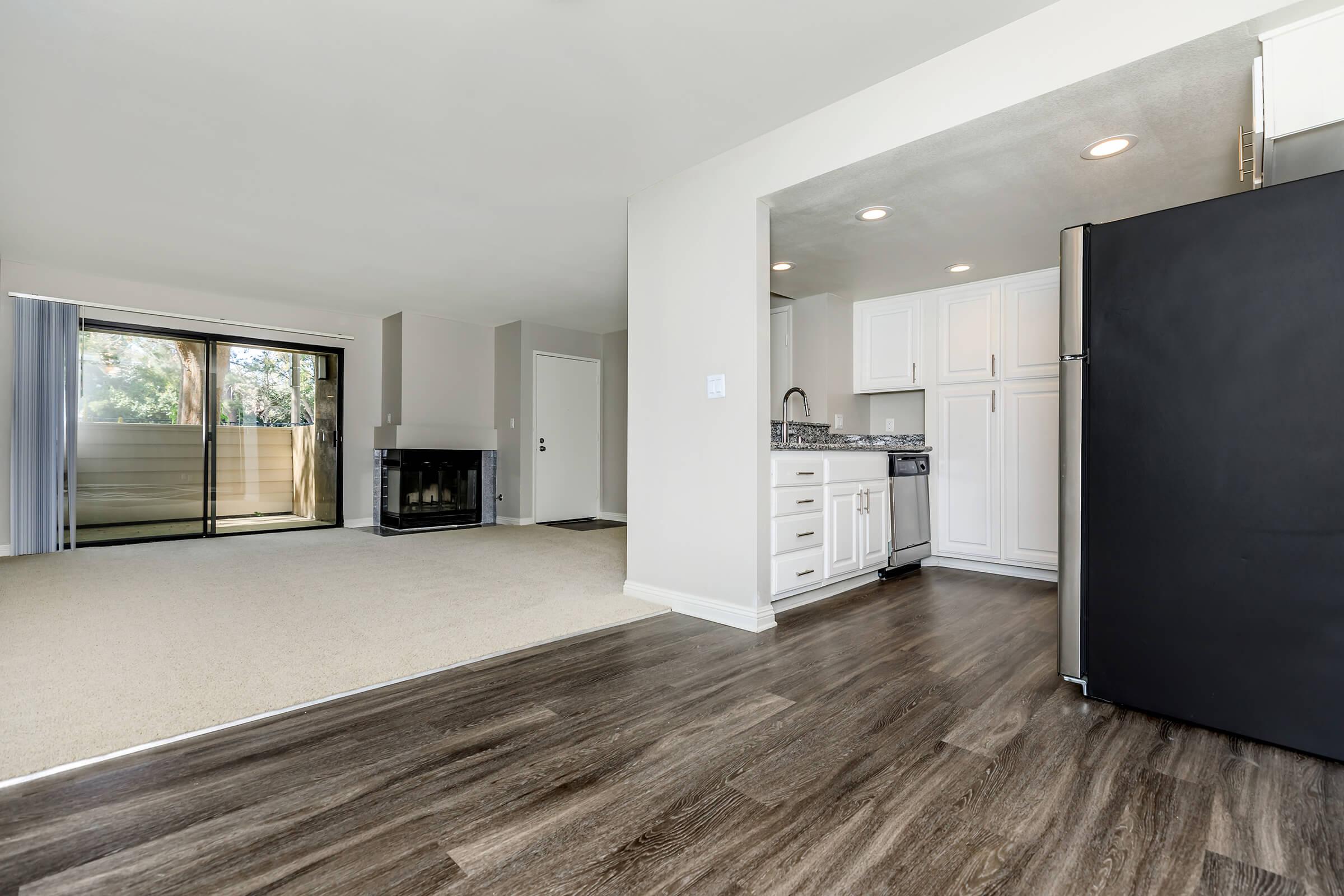 Spacious living area with a view of a balcony, featuring large windows for natural light. The kitchen is partially visible, equipped with white cabinets and modern appliances. The flooring is a combination of carpet and sleek wood, complemented by a cozy fireplace in the corner of the room.