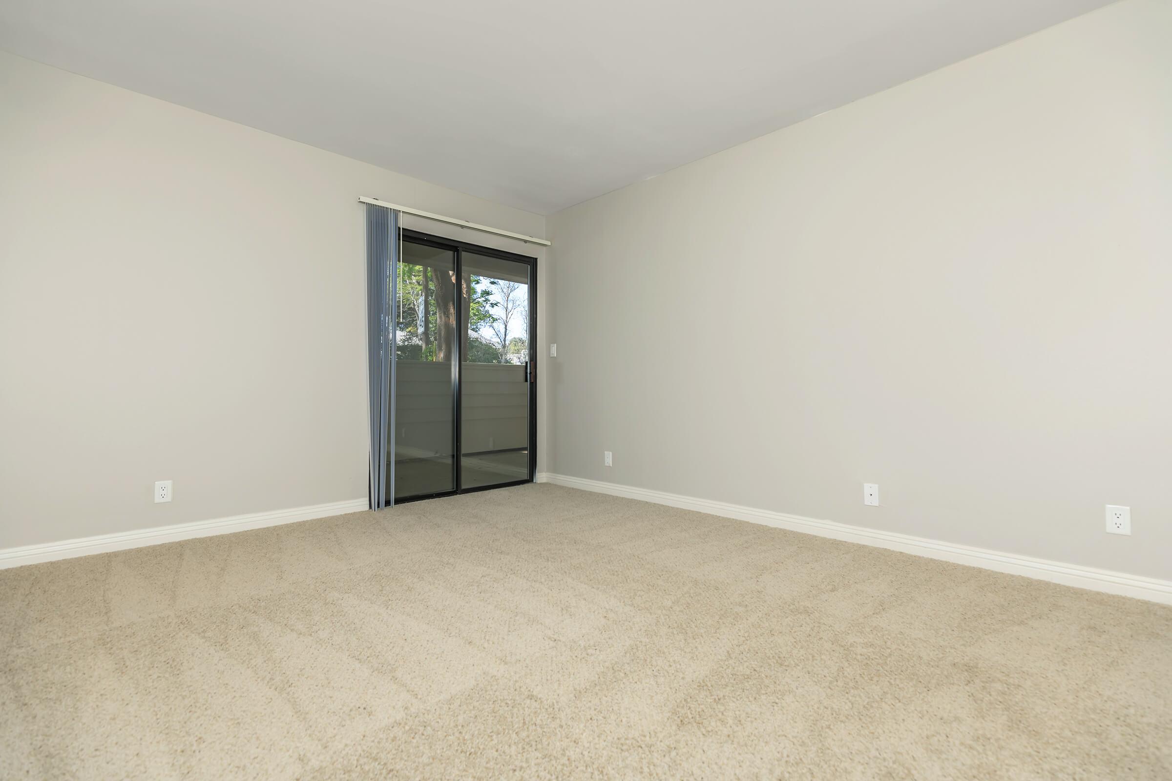 Empty room with light gray walls and a large window with sheer curtains providing natural light. The floor is covered in beige carpeting. A sliding glass door leads to a balcony, and there are electrical outlets on the walls. The space appears clean and ready for furnishing.