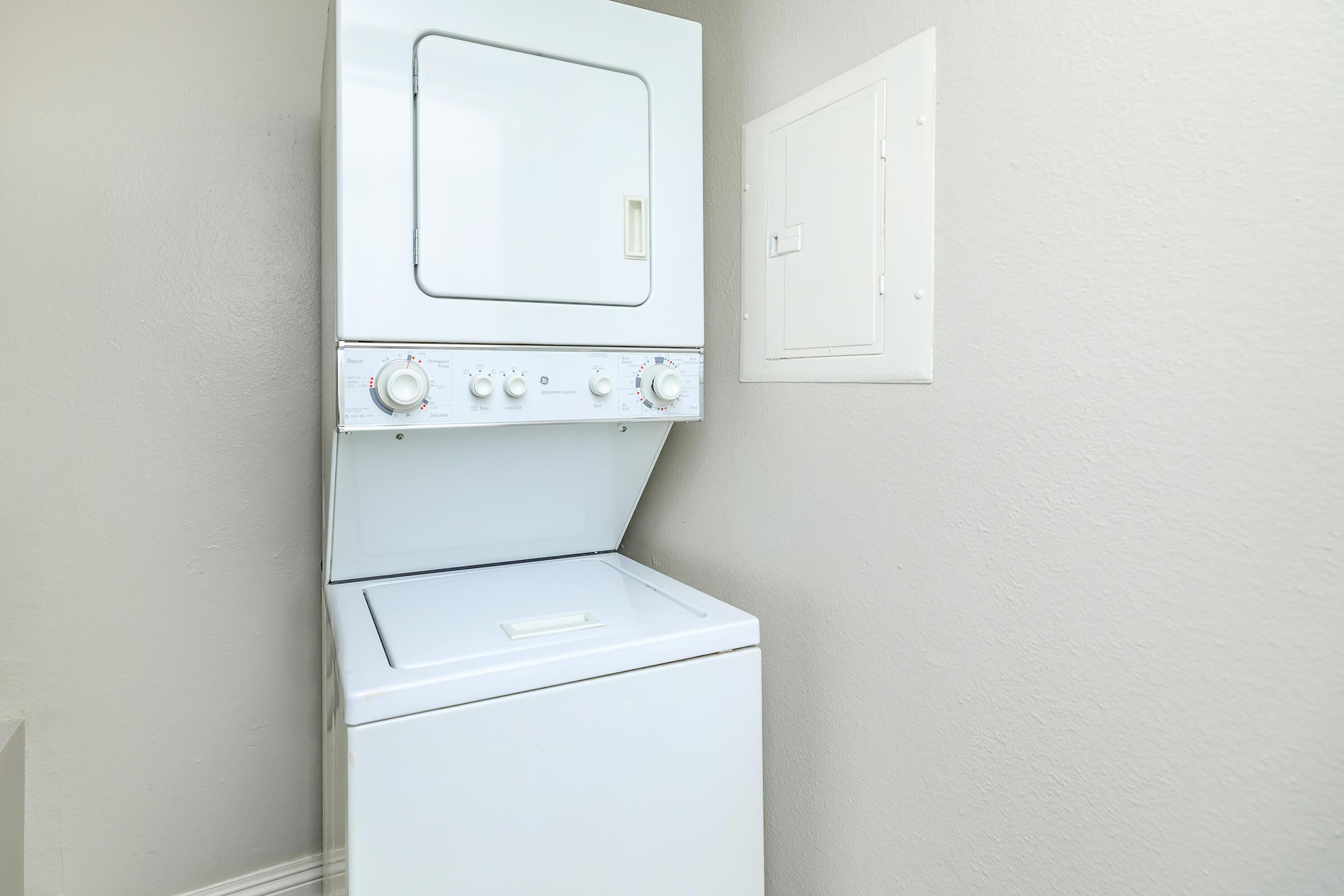 Stacked washer and dryer unit in a laundry corner, featuring controls for various wash settings. A small access panel is visible on the wall beside the machine, with neutral-colored walls enhancing the clean, functional look of the space.