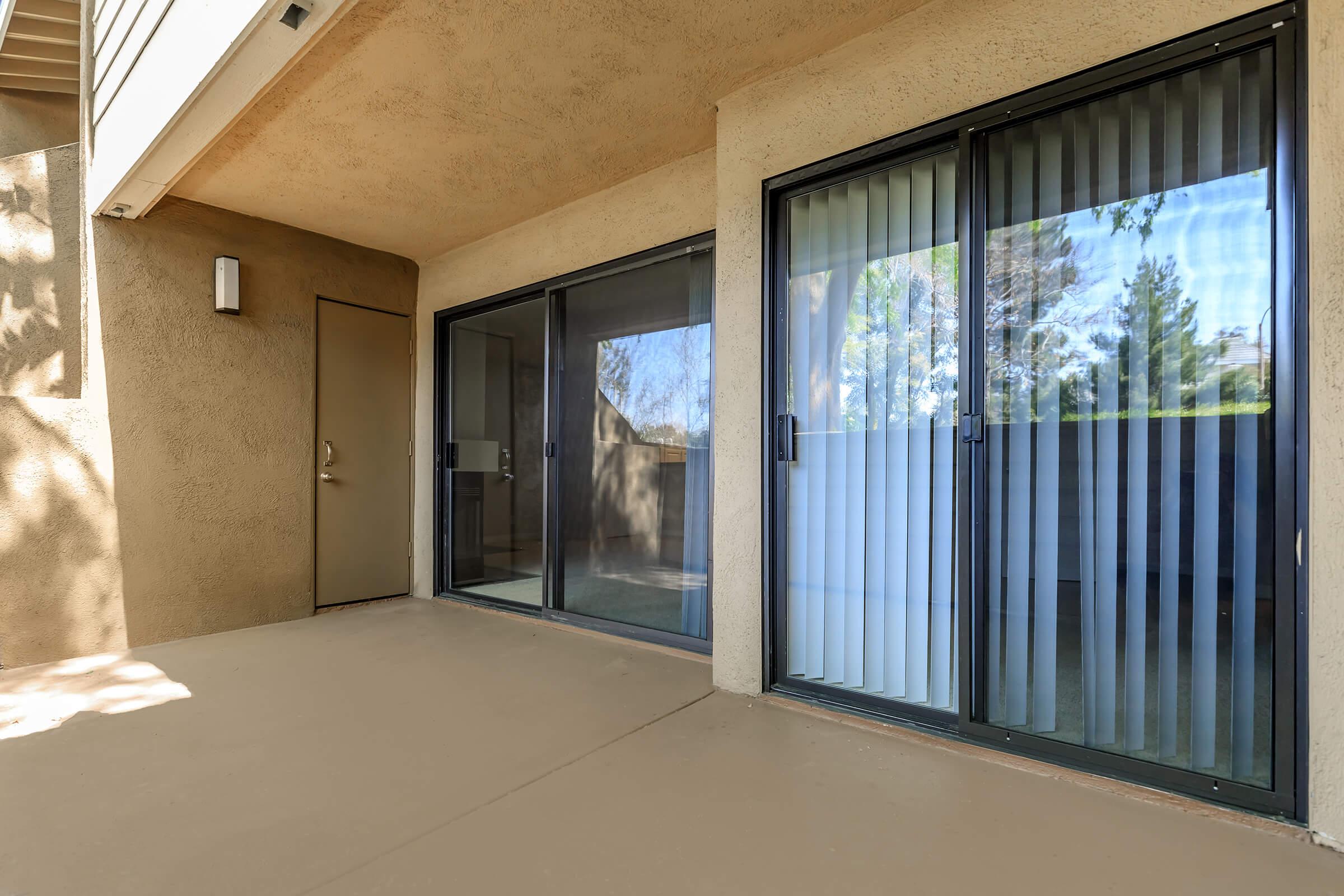 A patio area featuring a smooth concrete floor and two sets of sliding glass doors with vertical blinds. The walls are a light beige color, and there is natural light filtering in, suggesting a warm, inviting atmosphere. Trees are visible outside through the glass.