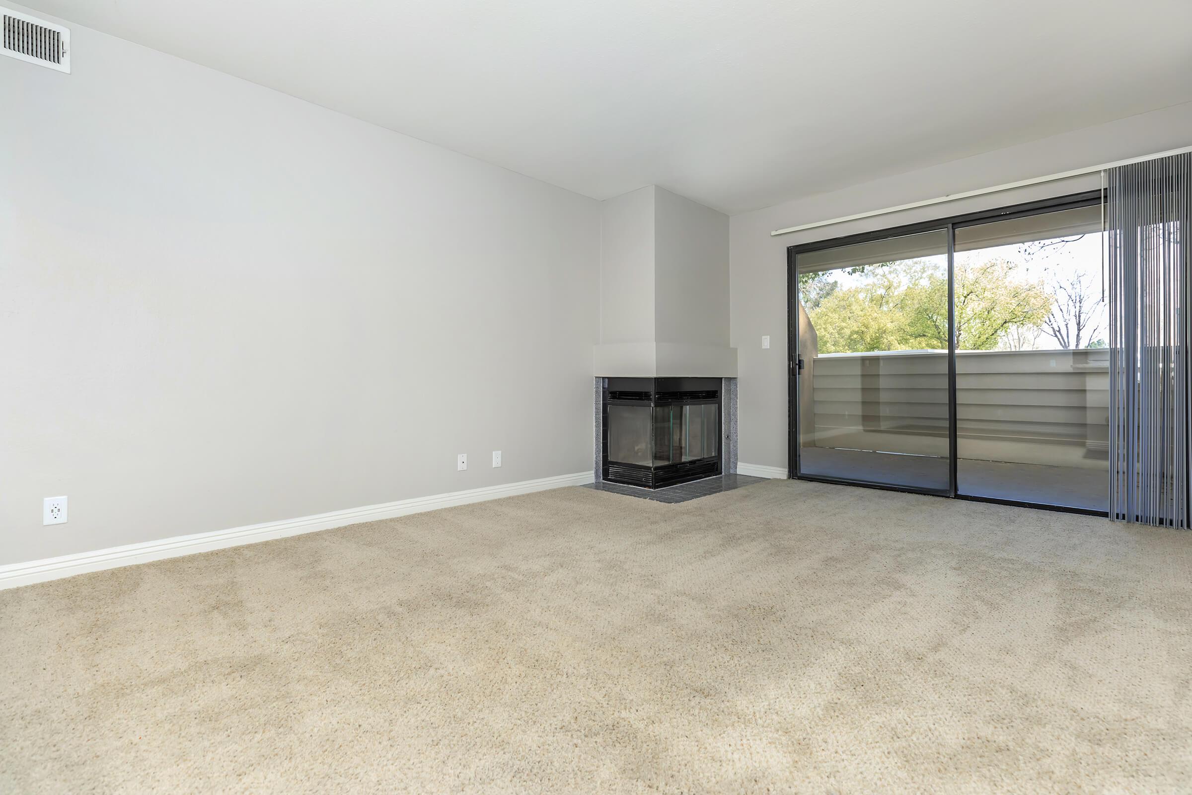 A spacious living room featuring a fireplace, light gray walls, and beige carpet. Large glass doors lead to a balcony with greenery visible outside. The room is well-lit and minimalistic, creating a cozy atmosphere.