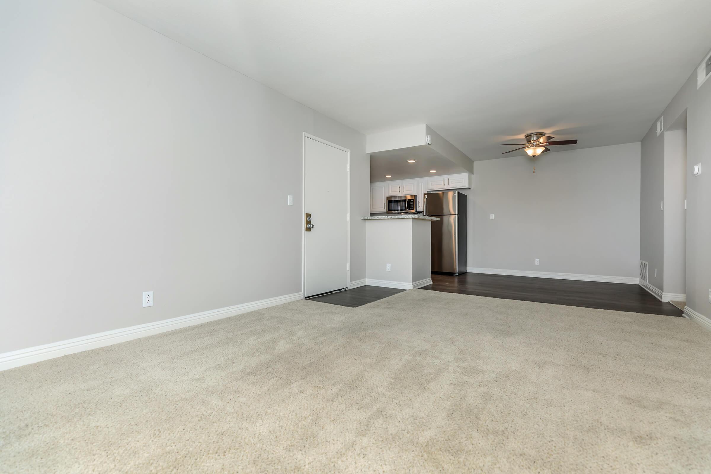 Empty living space with light gray walls and a tan carpet. A door is visible on the left side, leading to the outside. In the background, a kitchen area features stainless steel appliances and a ceiling fan above. The space is well-lit and modern, creating an inviting atmosphere.