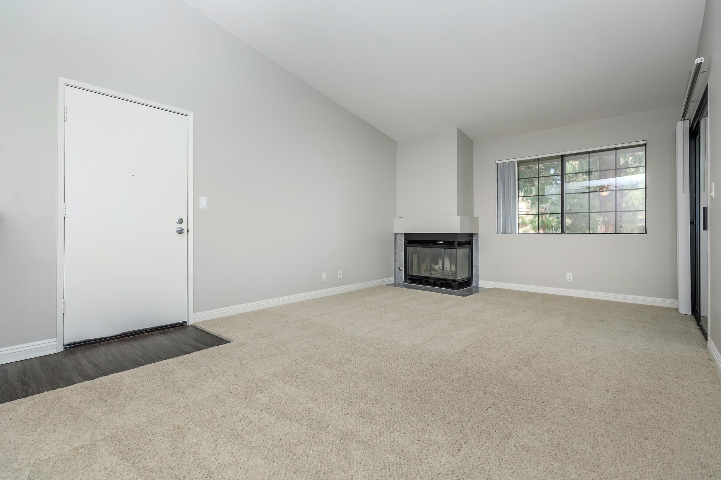A spacious and empty living room featuring light gray walls, a window providing natural light, a cozy fireplace in the corner, and soft beige carpeting. The entrance door is visible on the left, and the room has a minimalist vibe, making it suitable for personalization.