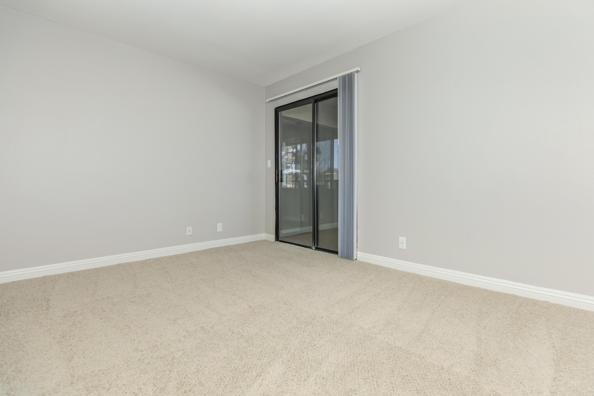 A spacious, empty room with light gray walls and beige carpet. There is a large glass door leading to an outdoor area, covered by sheer curtains, allowing natural light to enter. The room is devoid of furniture, creating a minimalistic and open atmosphere.