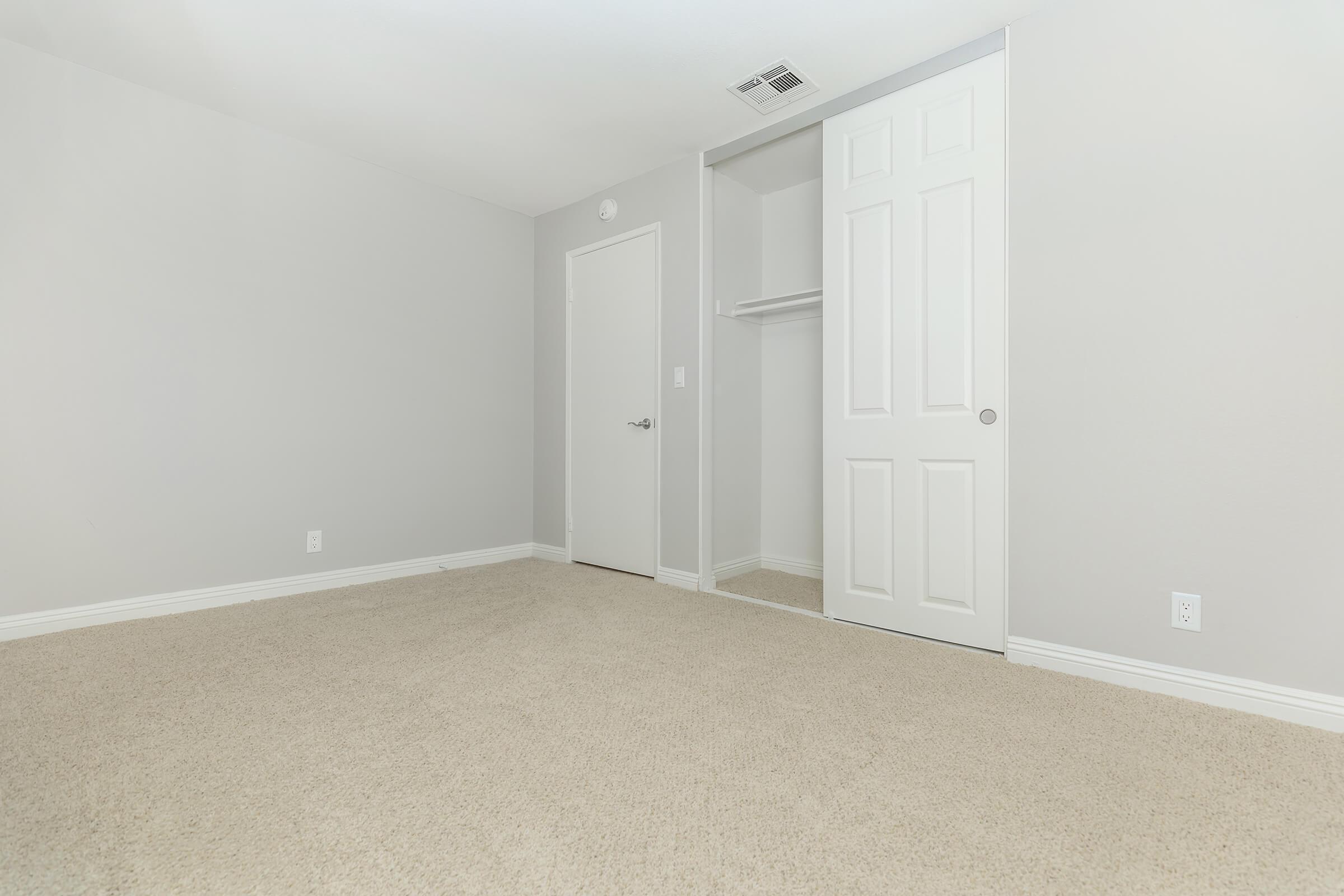 A spacious, empty room with light gray walls and beige carpet. It features a closed white door, a closet with mirrored doors, and no furniture or decorations, creating a blank canvas for potential design. Natural light illuminates the space, enhancing its openness.
