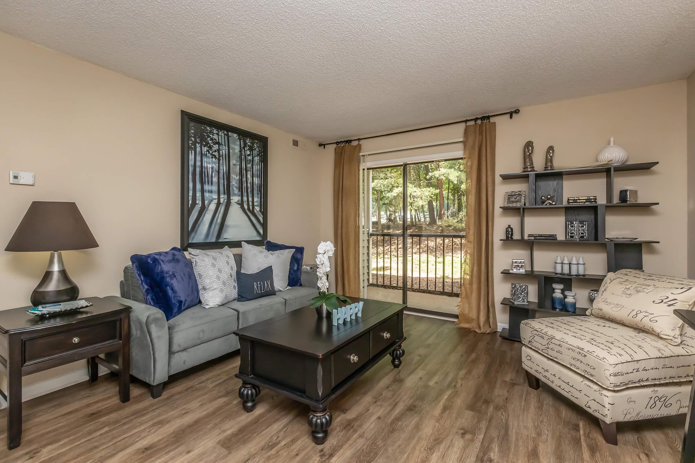 Cozy living room featuring a green sofa with blue and white pillows, a black coffee table, and a decorative lamp. A large window with sheer curtains provides natural light, overlooking a view of trees. A bookcase displays decor items, and a light-colored armchair complements the space.