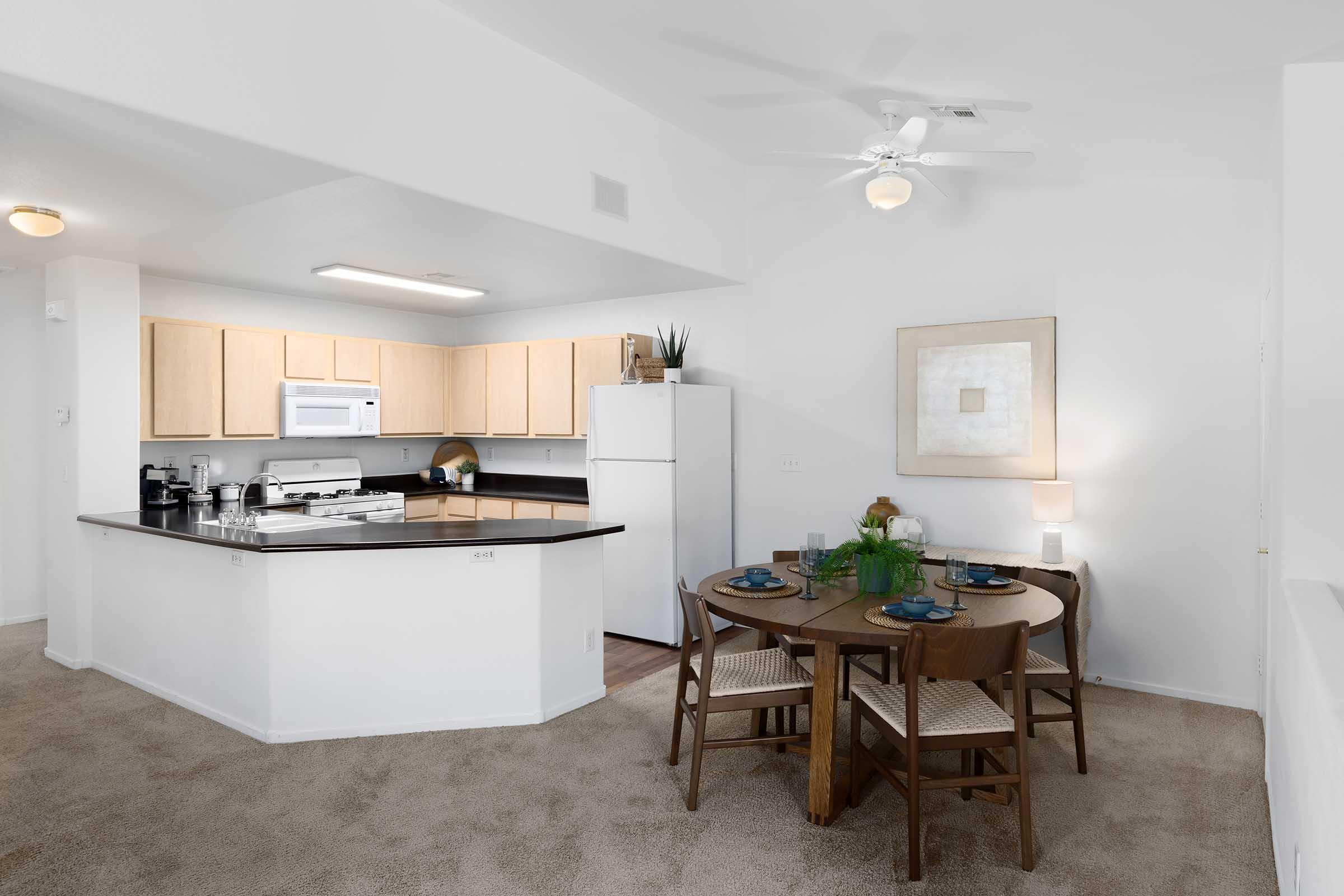 Modern kitchen and dining area featuring light wood cabinetry, a black countertop, and white appliances. A round dining table with four wooden chairs is set with dishes, while a ceiling fan and natural light enhance the bright, open space. Neutral carpet and decor create a cozy atmosphere.