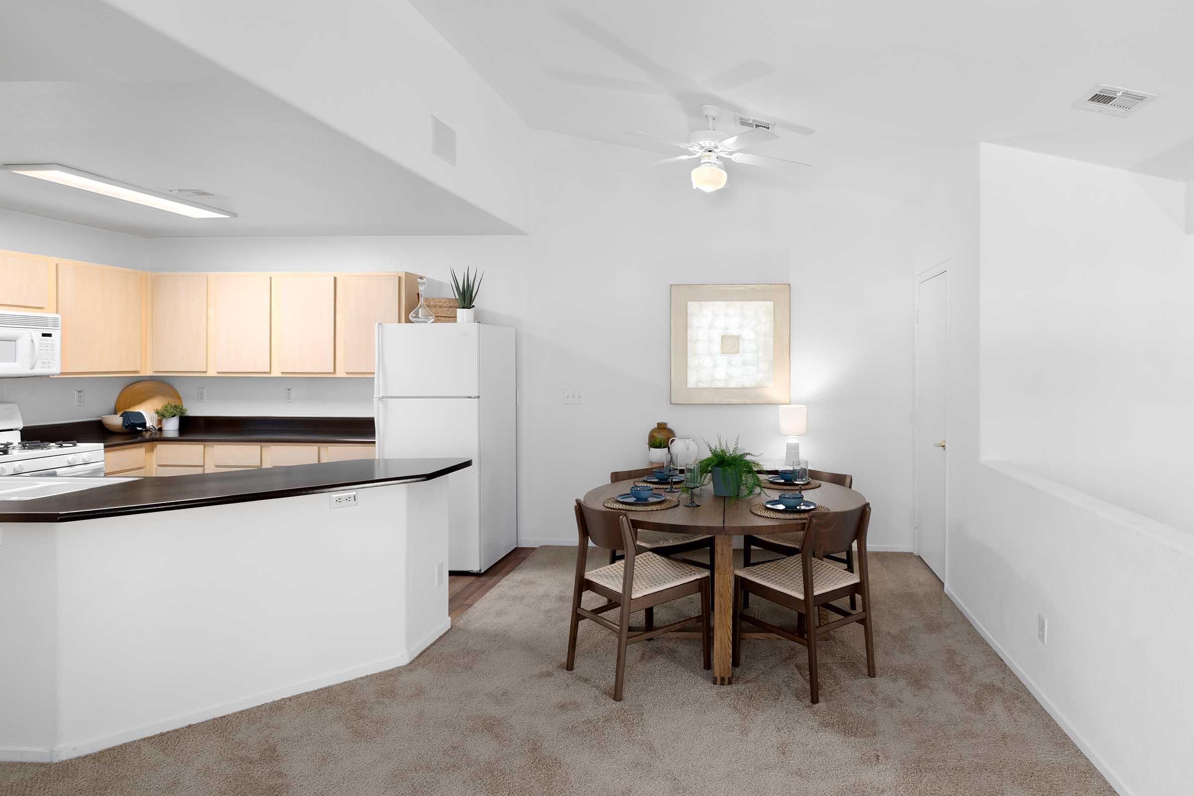 Cozy dining area featuring a round table with place settings, surrounded by four chairs. In the background, a well-equipped kitchen with light wood cabinets, a refrigerator, and a ceiling fan. Natural light fills the space, complemented by a simple piece of wall art and decorative plants.
