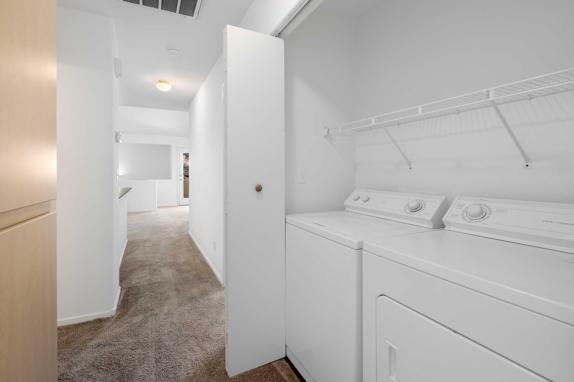 A view of a laundry room featuring a washing machine and dryer side by side, with a door partially open leading to a hallway. The room has beige carpeting and white walls, with a shelf above the appliances for storage. The hallway in the background is well-lit and leads to another area of the home.