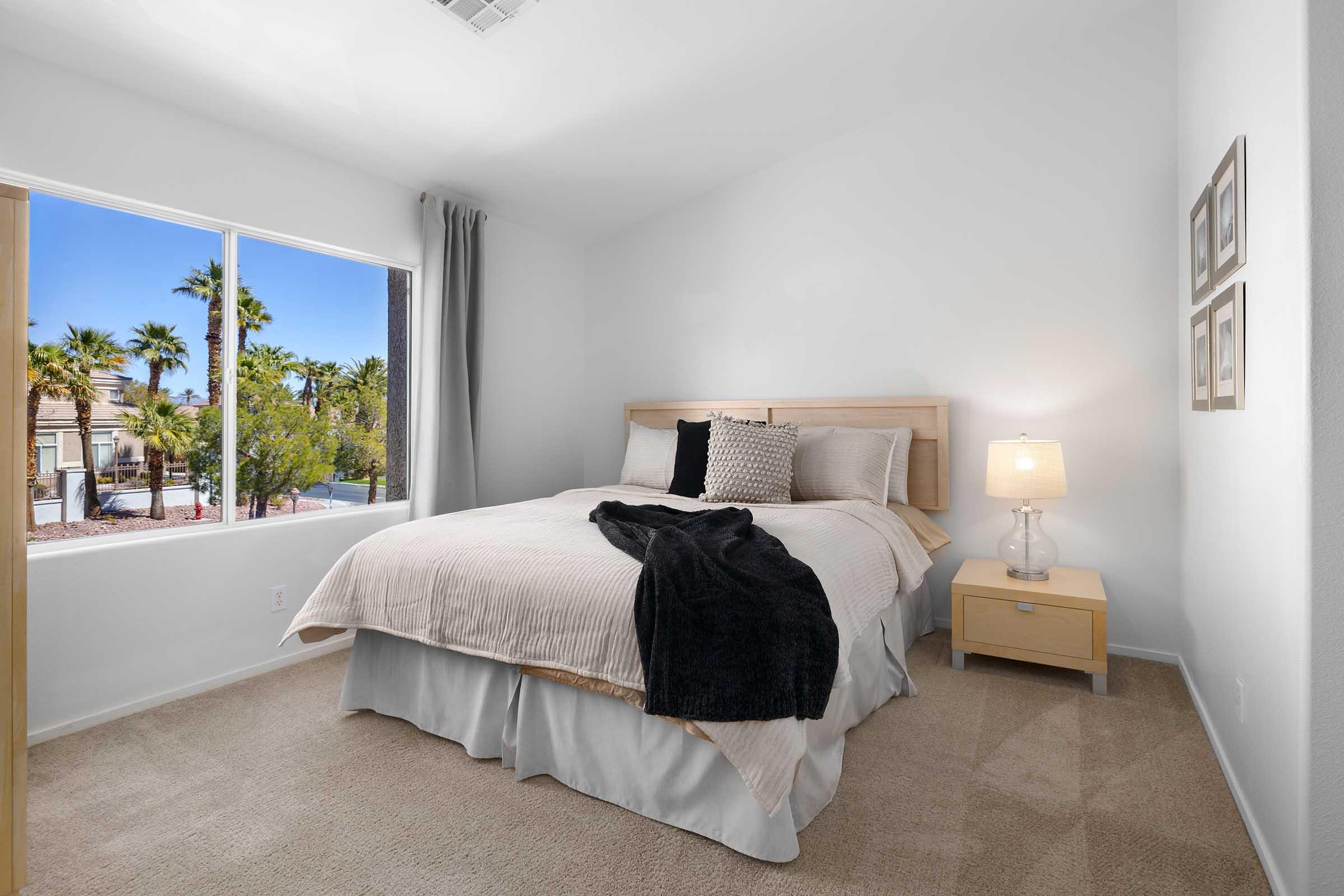 A cozy bedroom featuring a queen-sized bed with beige bedding, a black throw blanket, and a bedside table with a lamp. The room is bright with natural light coming from a large window, offering a view of palm trees outside. The walls are painted white, creating a clean and airy atmosphere.