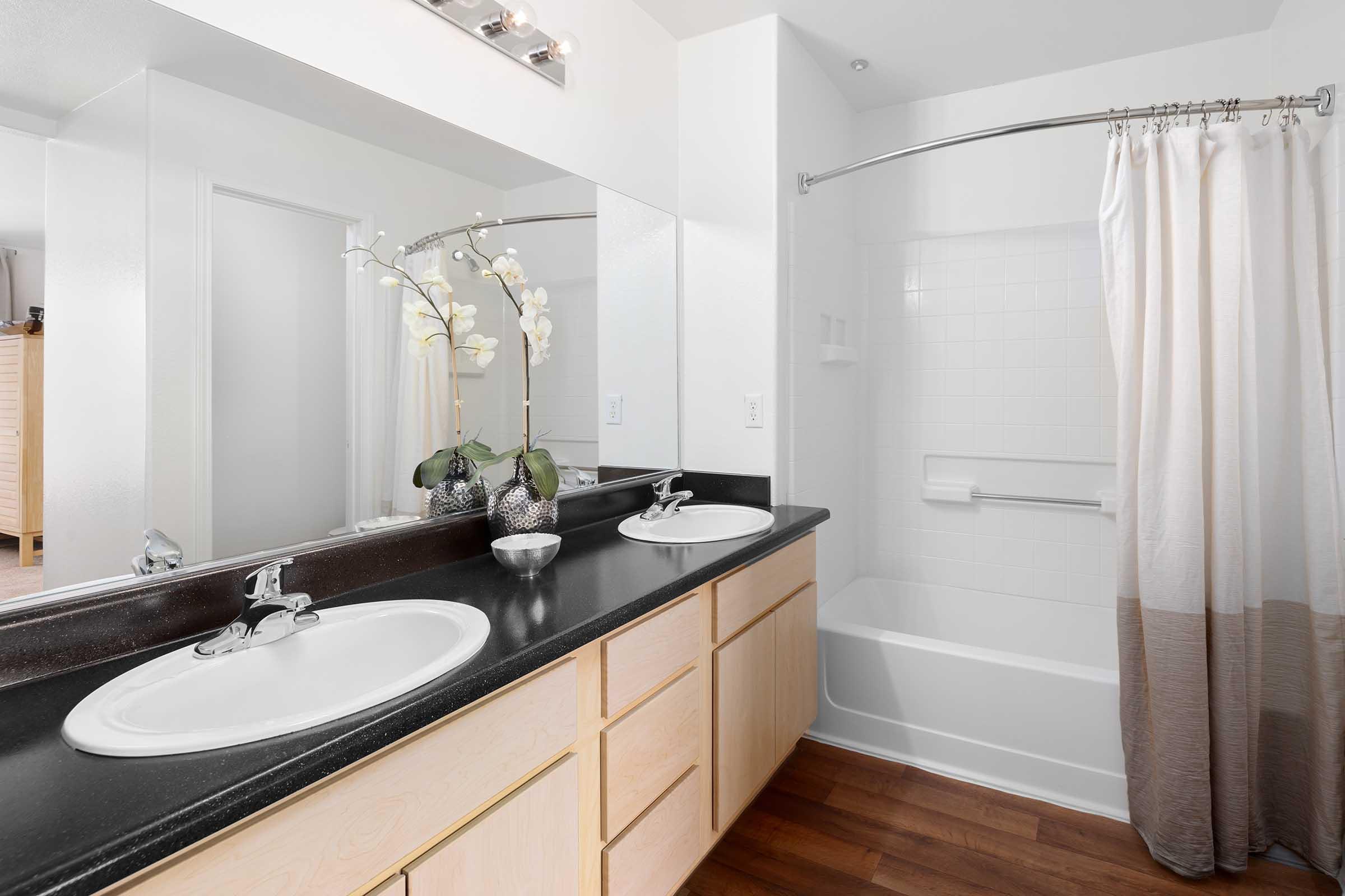 A modern bathroom featuring a double sink vanity with a dark countertop, two mirrors above the sinks, and a bathtub with a shower curtain. A decorative vase with orchids is placed on the counter. The walls are light-colored, and the flooring is wooden.
