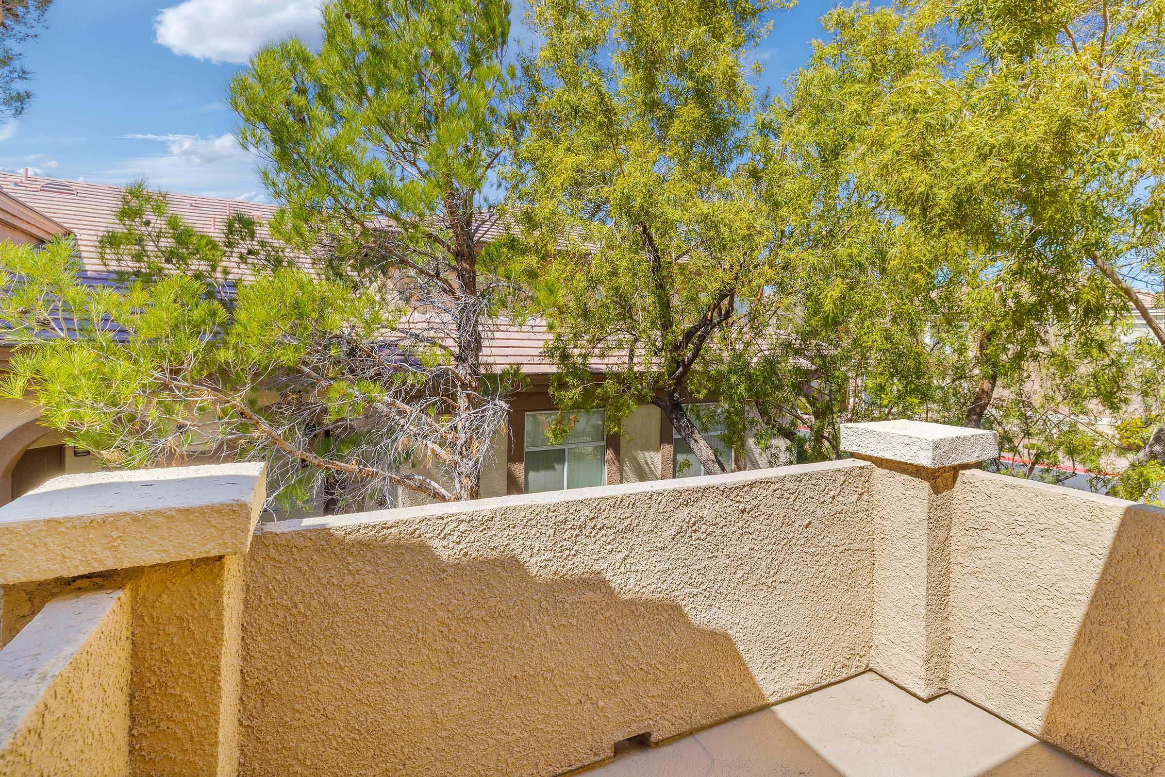 View from a balcony overlooking a tree-filled courtyard with residential buildings in the background. The image features a bright sky with scattered clouds, and the balcony is framed by stone railing and textured walls.