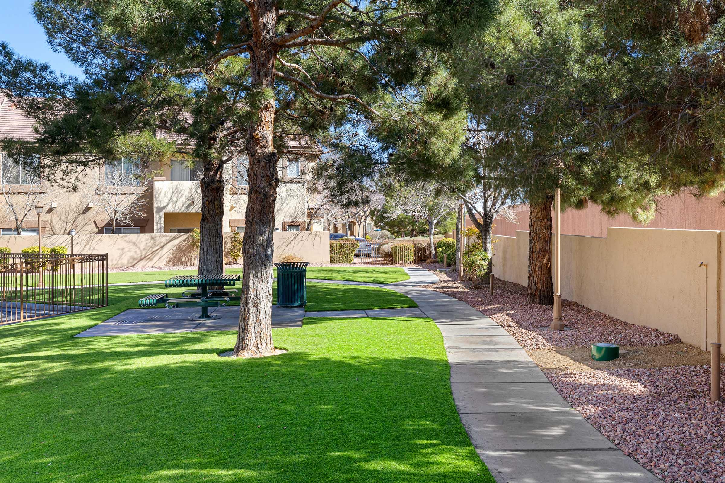 A well-maintained pathway lined with trees, leading through a green grassy area. On one side, there are picnic tables and a trash bin, while smooth concrete paths curve through the landscape. The background features buildings and a wall, contributing to a peaceful outdoor setting.