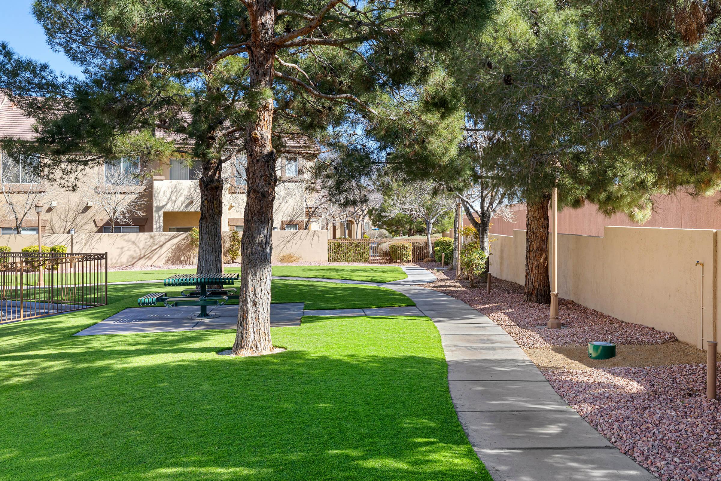 A well-maintained green outdoor space featuring a winding pathway bordered by trees. In the background, there are residential buildings. A fenced area with seating is visible, along with a patch of gravel and a small green bucket. The setting appears sunny and inviting, ideal for leisurely walks.