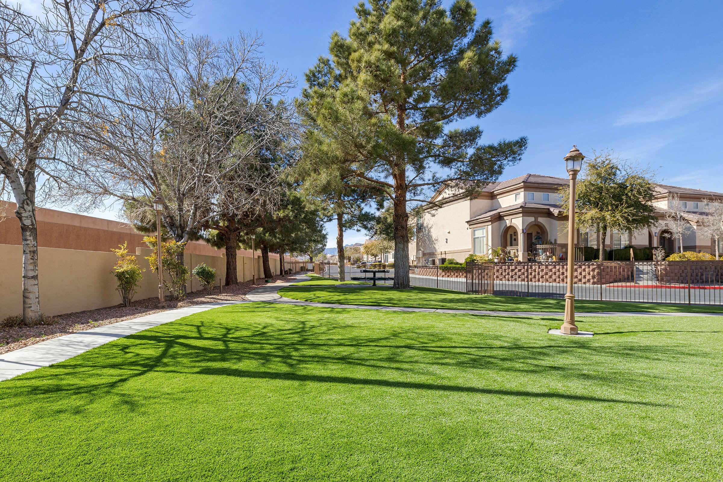 A well-maintained green lawn area featuring a pathway, trees, and a decorative lamp post. In the background, there are buildings with landscaped gardens. The scene is set under a clear blue sky, creating a pleasant outdoor space for relaxation or recreation.