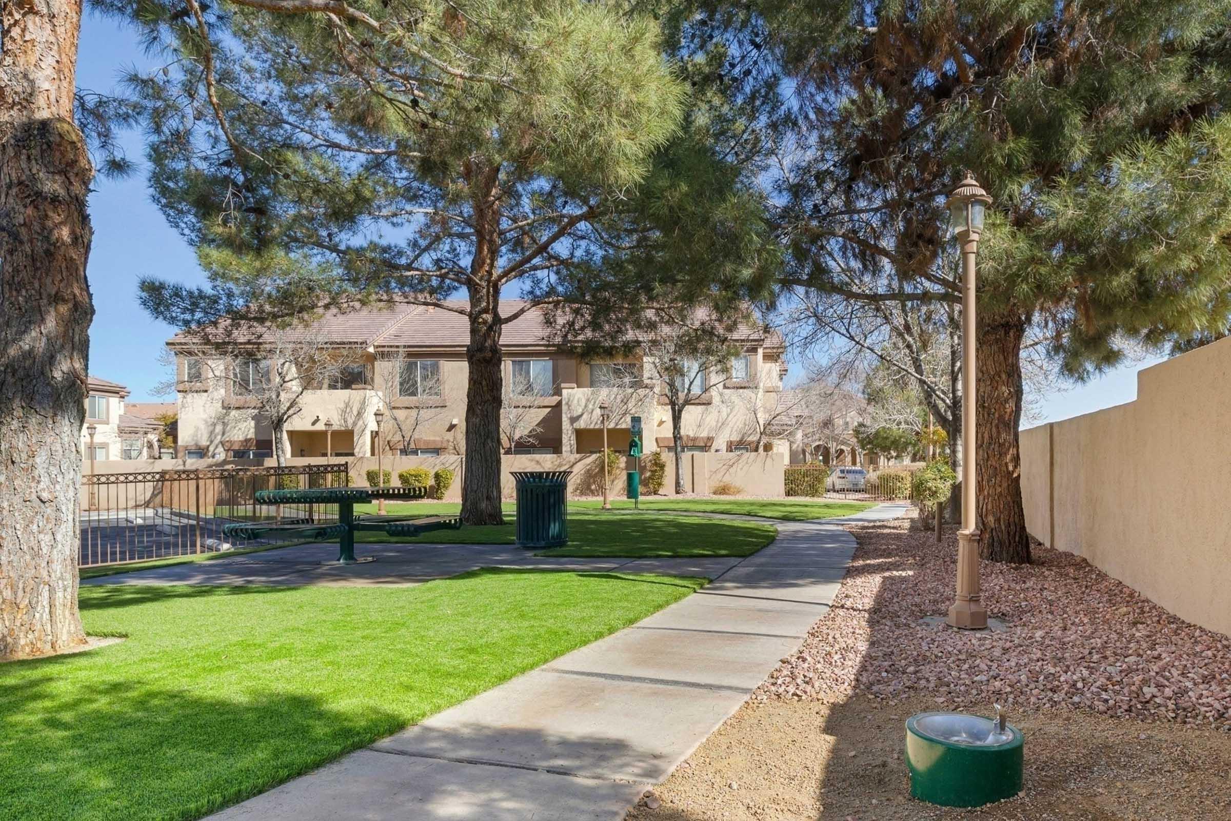 A community park area with a pathway lined by trees, featuring a green bench and trash bin. In the background, there are residential buildings. The scene shows manicured grass and gravel, creating a peaceful outdoor space.