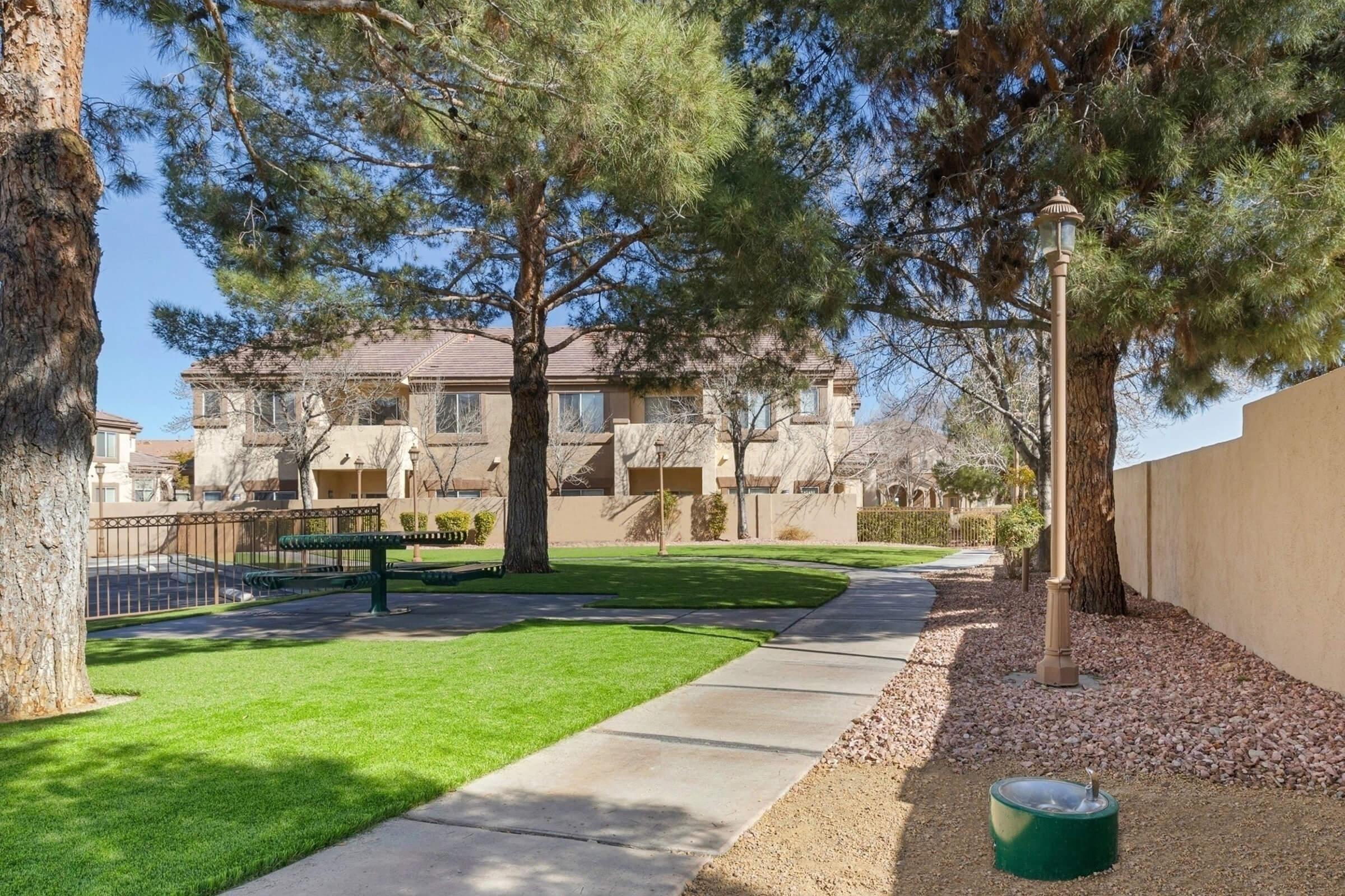 A landscaped area featuring well-maintained lawns, walking paths, and trees. In the background, there are two residential buildings with a fence. A bench is positioned near a pathway alongside the grassy area, creating a serene outdoor space.