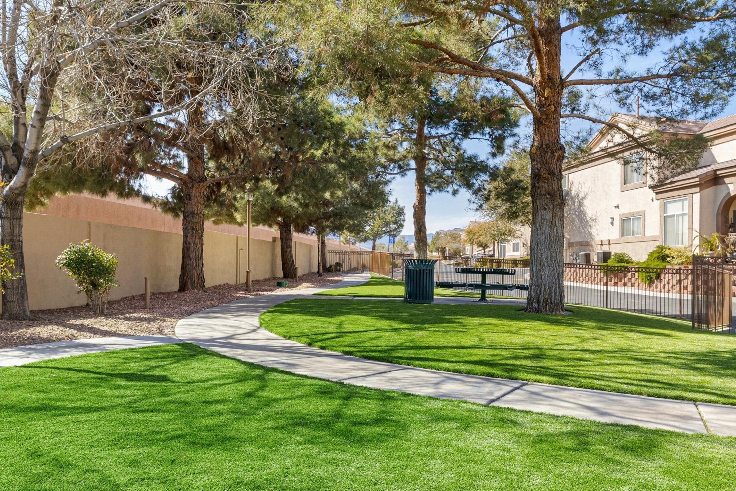 A peaceful park scene featuring a winding pathway surrounded by lush green grass, tall trees, and a trash bin. In the background, there are residential buildings with a low fence, creating a serene atmosphere perfect for walking or relaxing outdoors.
