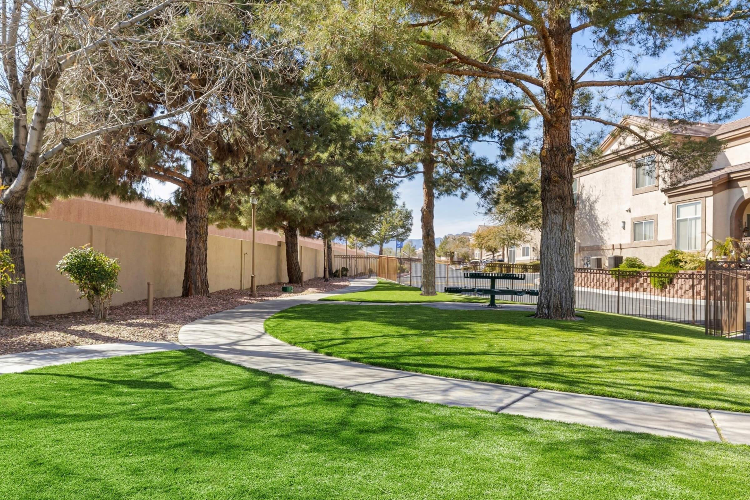 A well-maintained park area featuring a curved pathway lined with trees and green grass. A concrete bench and picnic table can be seen in the background, with a wall and residential buildings in the distance. Clear blue skies above, creating a serene outdoor environment.