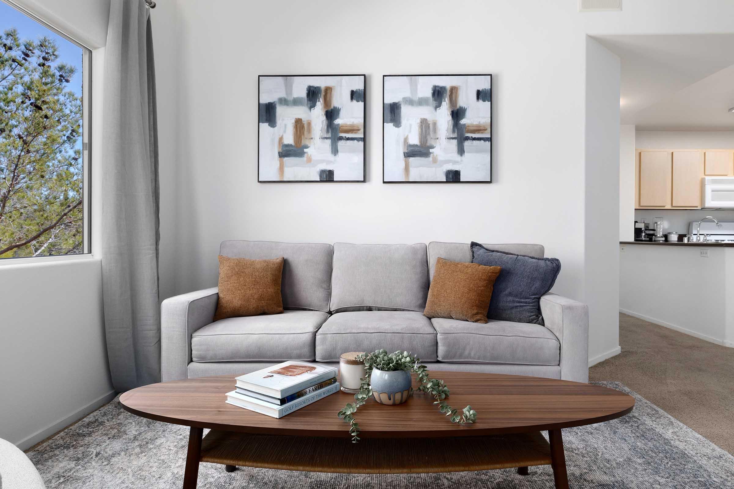 A cozy living room featuring a light gray sofa with decorative cushions, a wooden coffee table with books and a small plant, and two abstract wall art pieces. Natural light streams in through a window with sheer curtains, and a glimpse of a kitchen can be seen in the background.