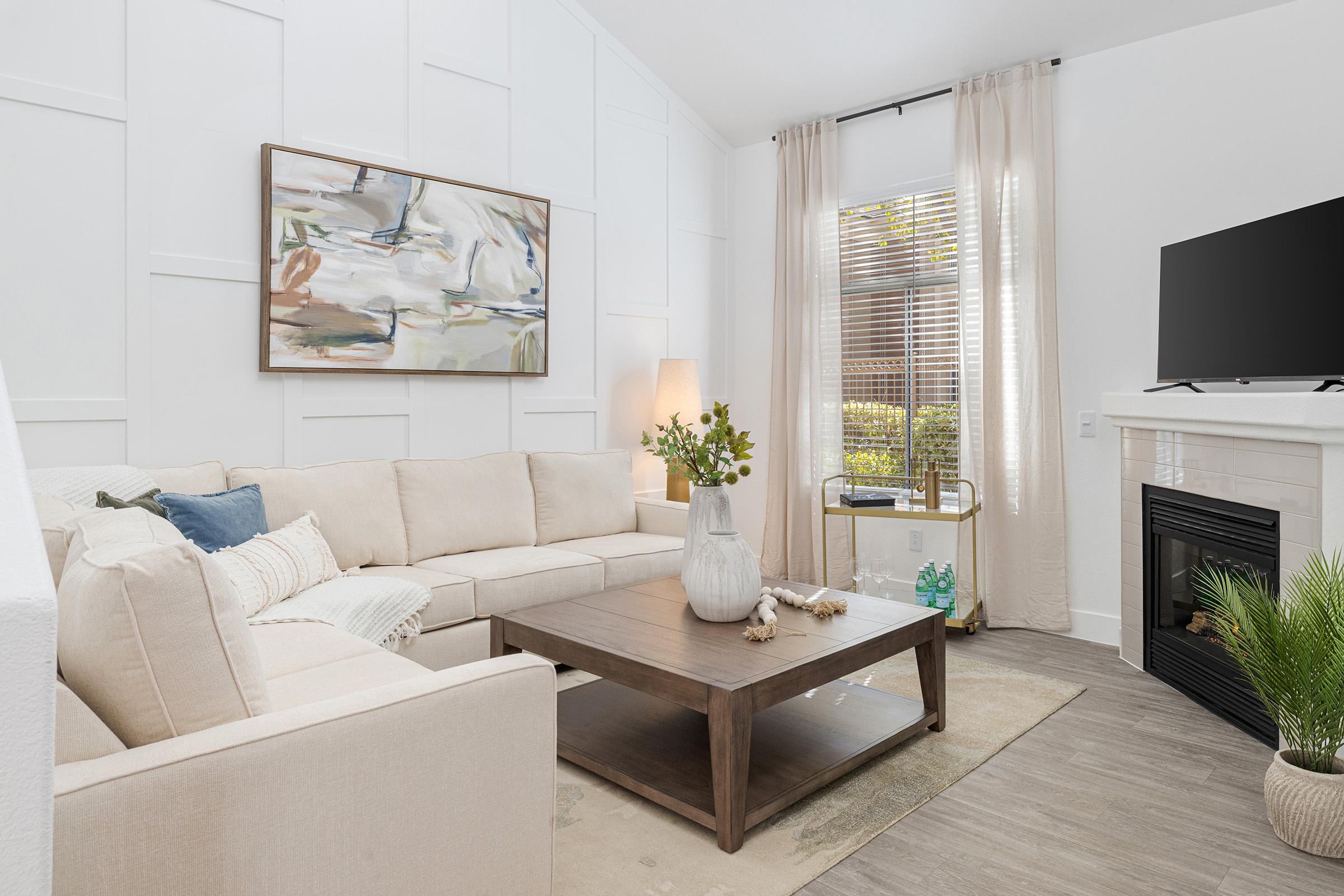 Bright living room featuring a light-colored sectional sofa, a wooden coffee table, and a wall-mounted TV. A piece of abstract art hangs above the sofa, and natural light pours in through large windows with sheer curtains. A decorative plant and a vase sit on the coffee table, adding a touch of greenery.