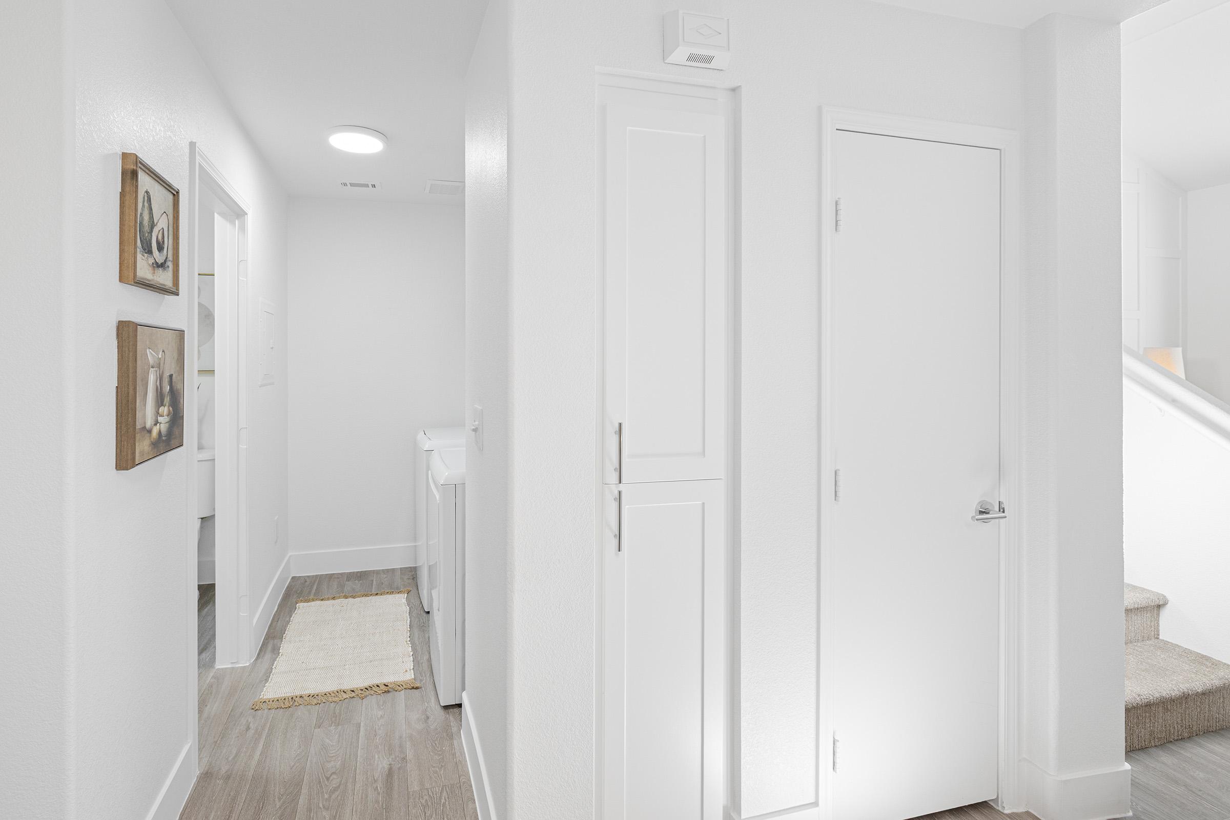 Bright, modern hallway featuring white walls and a light wood floor. On the left, there are two framed art pieces near a laundry area. To the right, there's a closed door and a small closet. A staircase is visible at the end of the hallway, leading to an upper level, illuminated by soft overhead lighting.
