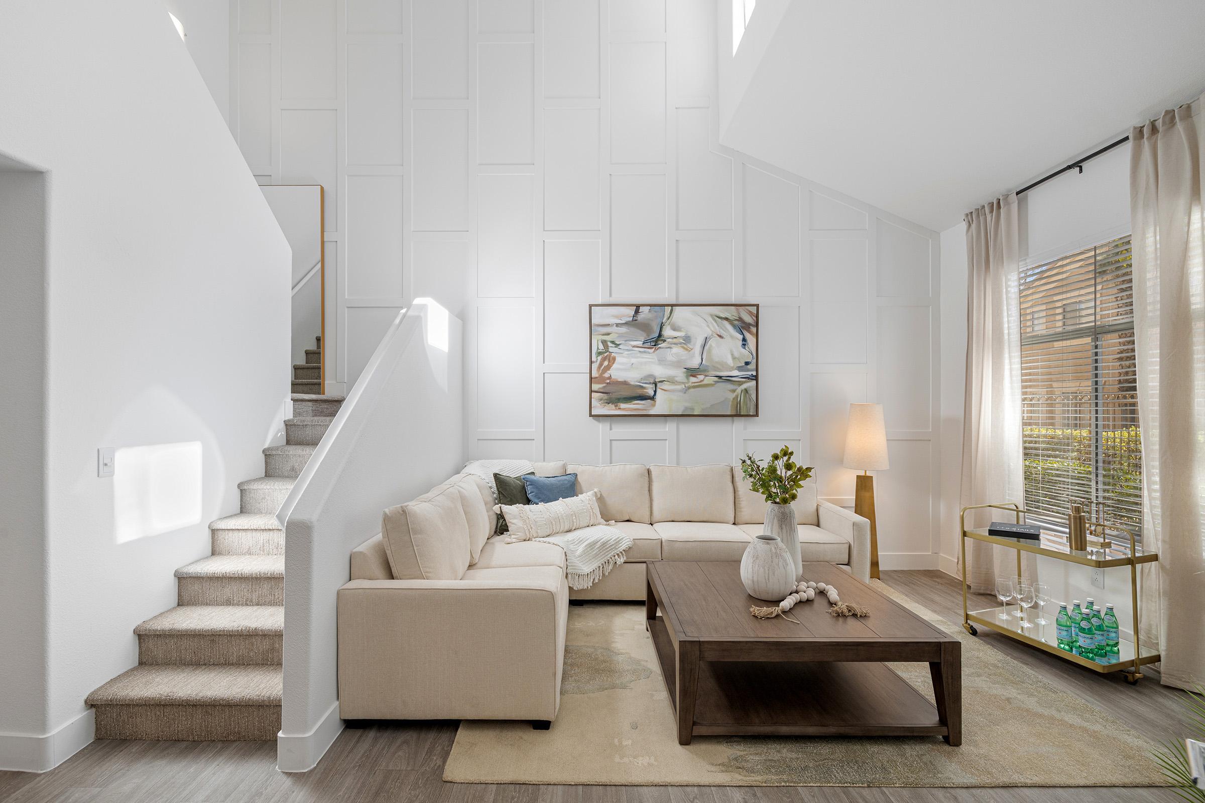 Bright and modern living room with a large cream sectional sofa, a stylish wooden coffee table, and a decorative vase. A staircase leads to an upper level, and a piece of abstract art hangs on the wall. Natural light filters in through sheer curtains, creating a warm and inviting atmosphere.