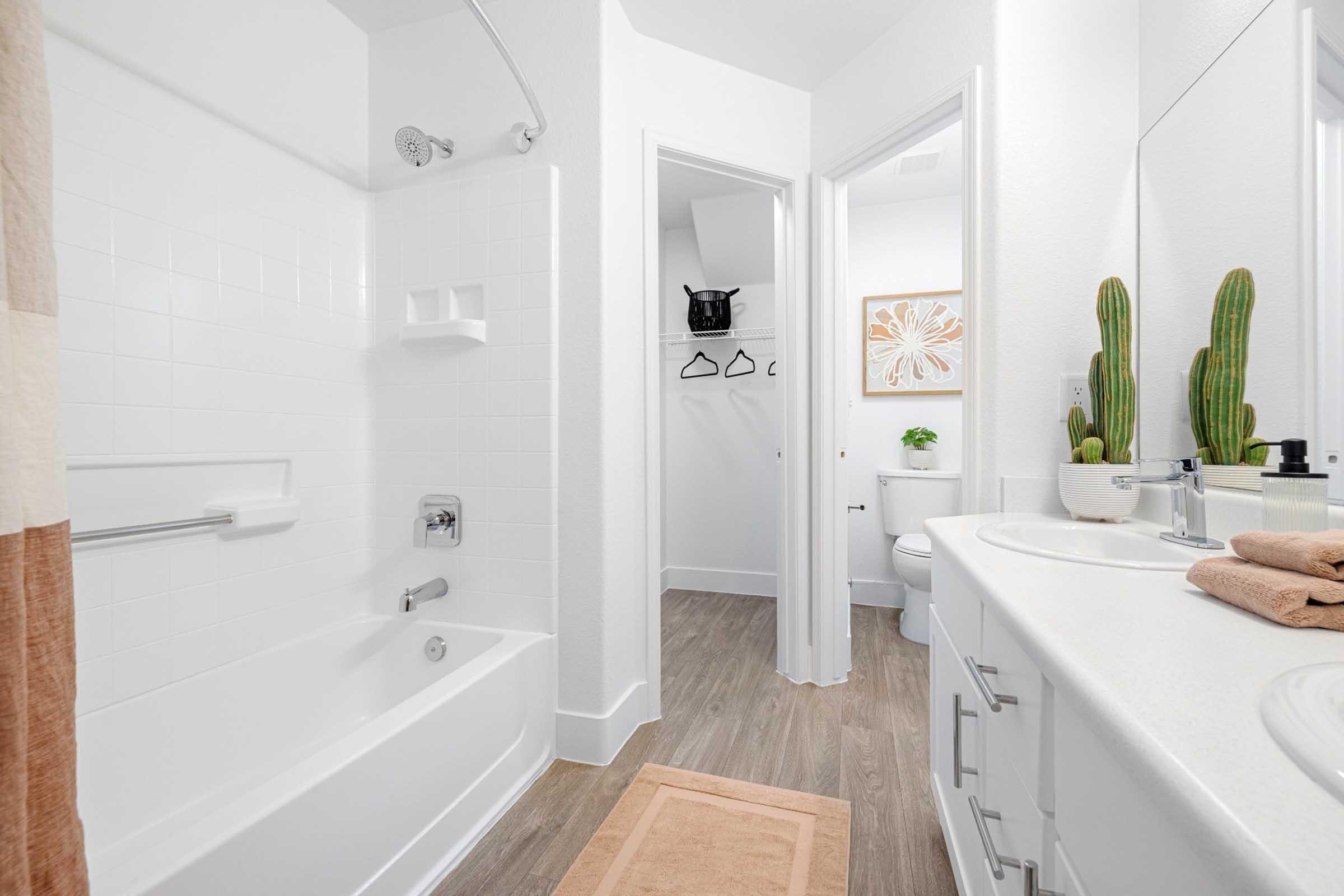 A modern bathroom featuring a white bathtub with a shower, a large mirror above a dual sink vanity, and light-colored walls. Cactus plants add a touch of greenery, while a tan bath mat is placed in front of the sink. A door leads to a small closet or storage area, and another door opens to a separate toilet area.