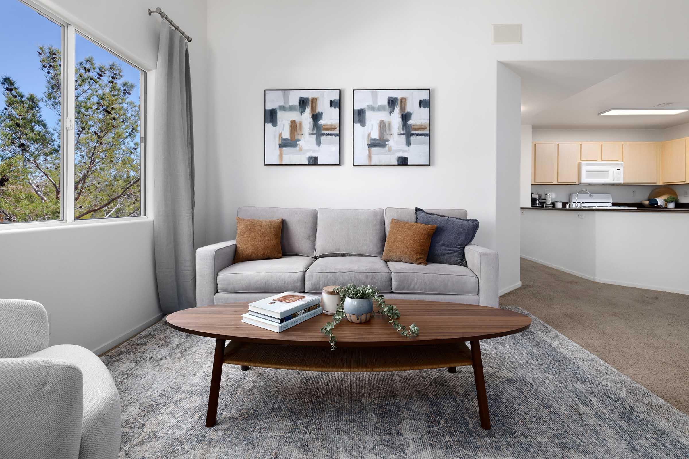 A modern living room featuring a grey couch with two brown pillows, a wooden coffee table with decor items, and two framed abstract paintings on the wall. Sunlight filters through a large window, illuminating the cozy space, while a doorway leads to a kitchen in the background.